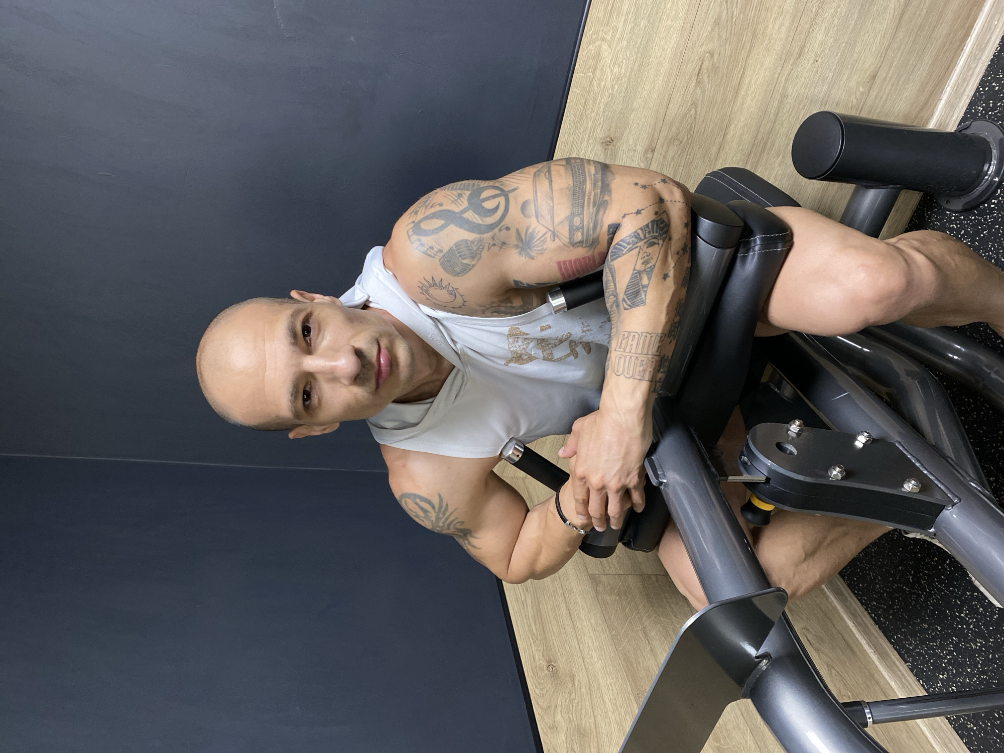 The image depicts a muscular, bald man with numerous tattoos on his arms and shoulders, likely within a gym setting. The man is seated on an exercise machine with black padding and gray metallic components, gripping the machine’s handles. The floor appears to be wooden near the man and transitions to a black, speckled, rubber surface further away. A portion of a dark gray or black wall is visible in the background. Given the provided context, the location is the city of Pleven, Bulgaria.