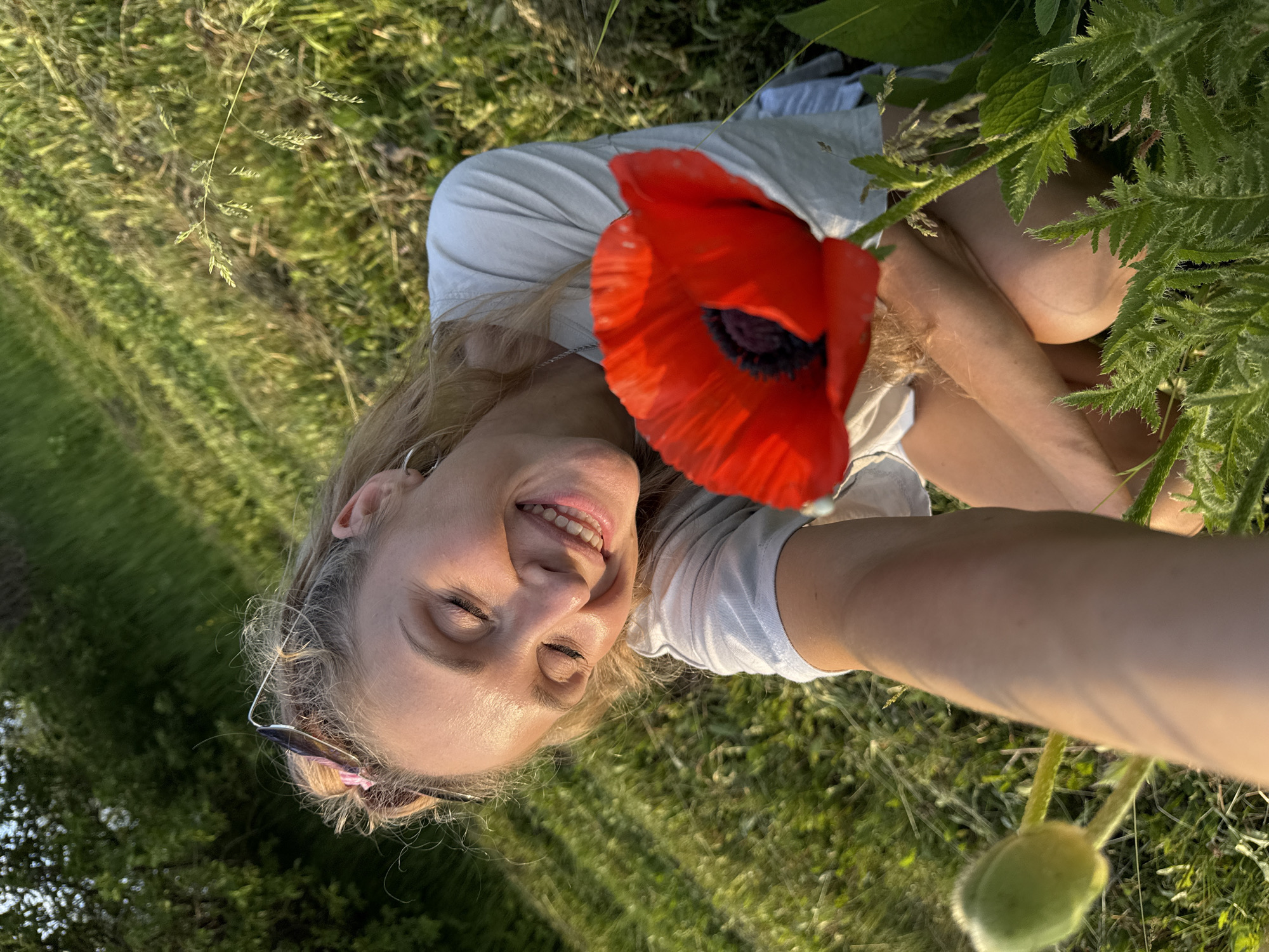 In a selfie taken outdoors in Nizhnyaya Chermoda, Russia, a smiling woman lies in a field of grass and wildflowers. She is wearing a light-colored t-shirt. A bright red poppy with a dark center is held up close to her face, partially obscuring it. Her eyes are closed, and her head is tilted back, suggesting a moment of enjoyment or relaxation. Sunglasses are perched on her head. The lighting suggests it is late afternoon or early evening, with the sun casting long shadows and illuminating the scene with a warm glow. The background is filled with lush green grass and other plants, indicating a natural, possibly rural, setting. The overall mood is peaceful and joyful. No visible text is present in the image.