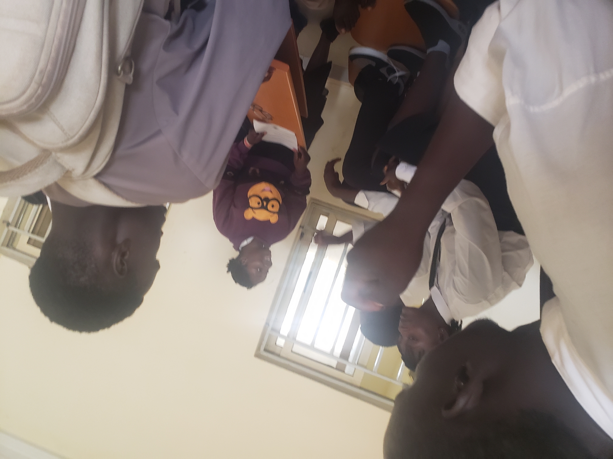 The image depicts a group of young people gathered in what appears to be a classroom or meeting room in Gumel, Nigeria. In the foreground, a person wearing a white shirt is partially visible on the left, with their back to the camera. To their right, two individuals in white shirts with dark ties are seated, facing each other and engaged in conversation. One of them is looking away from the camera. Further to the right, a woman wearing a purple sweatshirt with a character graphic and glasses is standing and holding a folder. She appears to be interacting with the seated individuals. In the background, a window with blinds and a doorway are visible. Another person, seen from behind, is wearing a light purple shirt and a light-colored backpack, with their head turned away from the camera. The environment suggests a casual or academic gathering.