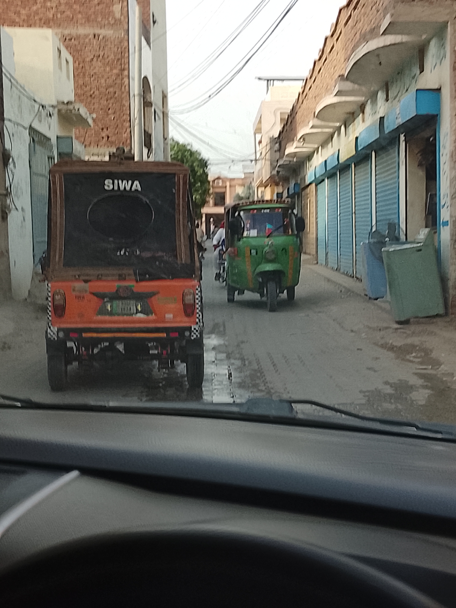 The image, captured from inside a vehicle, depicts a street scene in Nasheman Colony, Pakistan. Two auto-rickshaws, also known as tuk-tuks, dominate the view. The one closest to the camera is orange with "SIWA" displayed on the back. The second tuk-tuk is green. The image is taken from inside of a car.
Buildings with closed roll-up doors line the right side of the street.
Pedestrians are visible in the background.