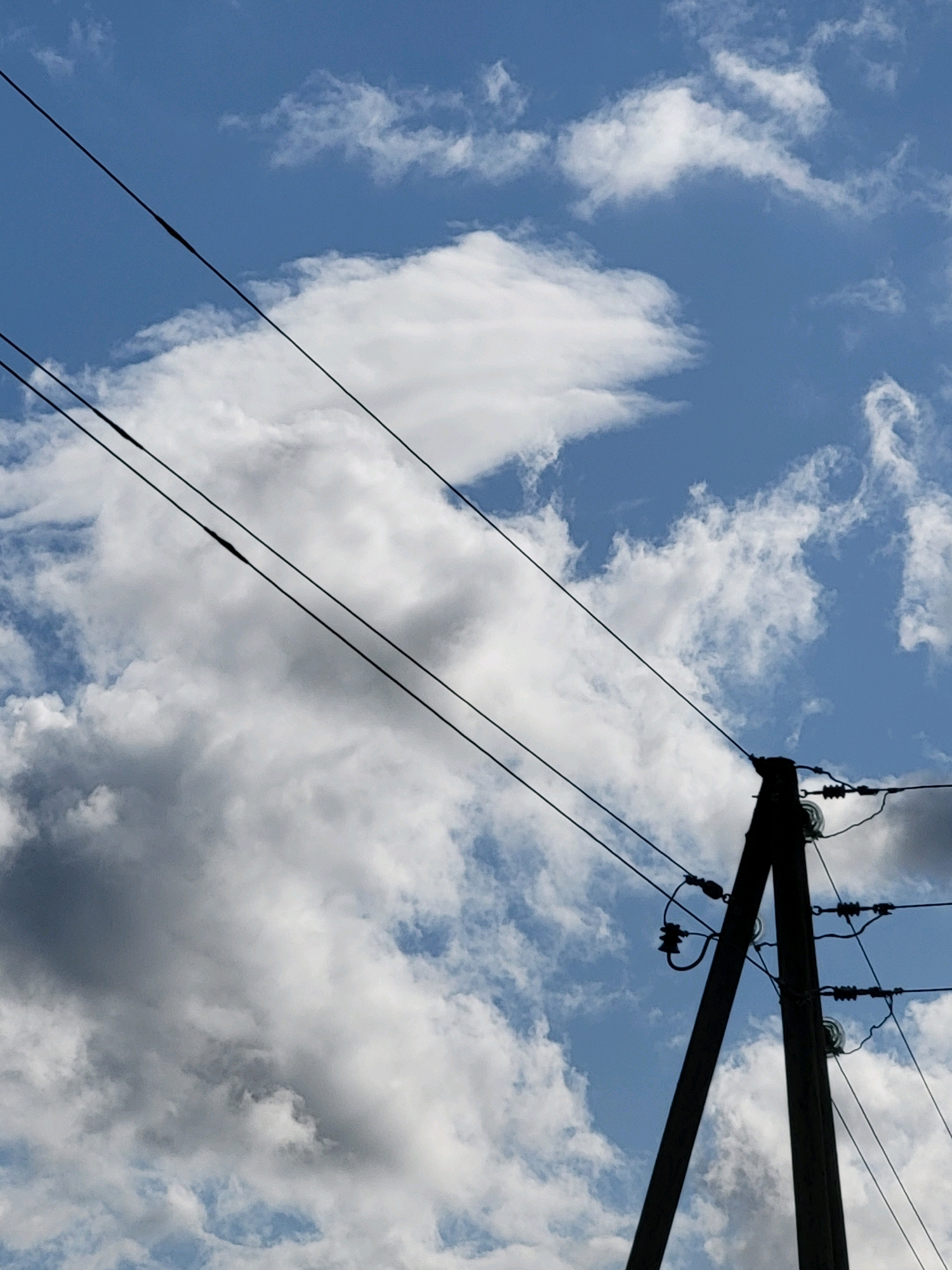 A low-angle, close-up shot captures a wooden utility pole silhouetted against a bright blue sky with scattered white clouds. The pole is tilted slightly to the right, with two diagonal crossbars extending to the left and right. Several wires are strung between the crossbars and extend upwards and to the left, disappearing out of frame. One wire in the foreground has a visible splice. The pole itself is dark and weathered, with insulators visible where the wires connect. The overall impression is one of an ordinary utility infrastructure against a dynamic sky, likely taken in an outdoor setting within the city of Jelgava, Latvia, given the context provided. There are no people or animals visible in the image.