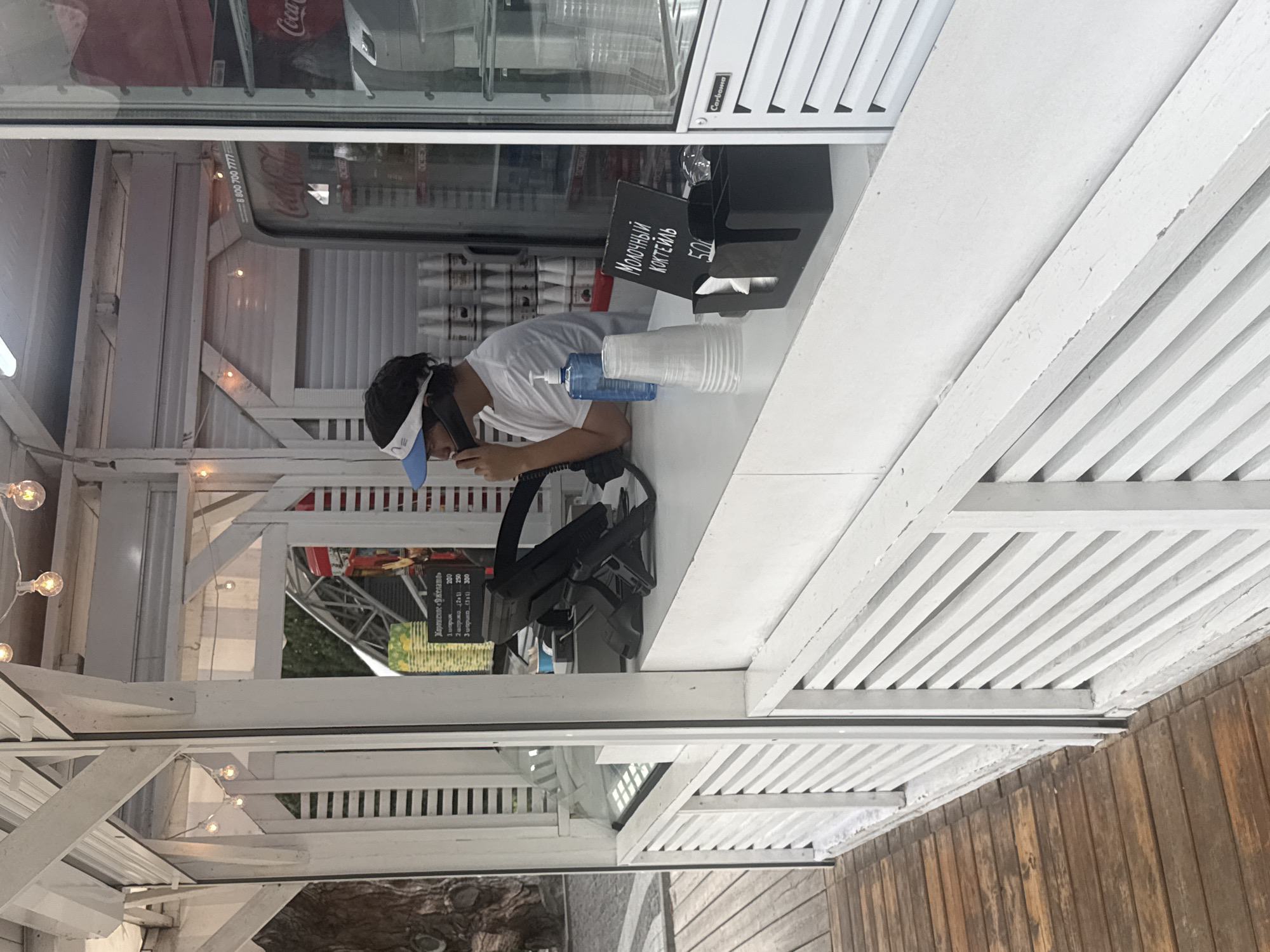 A young man wearing a white t-shirt and a blue and white visor is standing behind a white counter in an outdoor kiosk. He is holding a phone to his ear and appears to be speaking. On the counter, there are stacks of clear plastic cups, a blue dispenser of liquid, a black cash register, and a small black sign with white lettering that reads "МОЛОЧНЫЙ КОКТЕИЛЬ 50." Behind the counter, a Coca-Cola refrigerator is visible, stocked with drinks. The kiosk is made of white wood with slatted panels. The floor in front of the kiosk is made of wooden planks. The scene appears to be in an outdoor urban setting, possibly a promenade or park, given the wooden structures and implied public space. The time of day is daytime, with visible lighting fixtures on the kiosk. The location is identified as the city of Yalta, Ukraine.