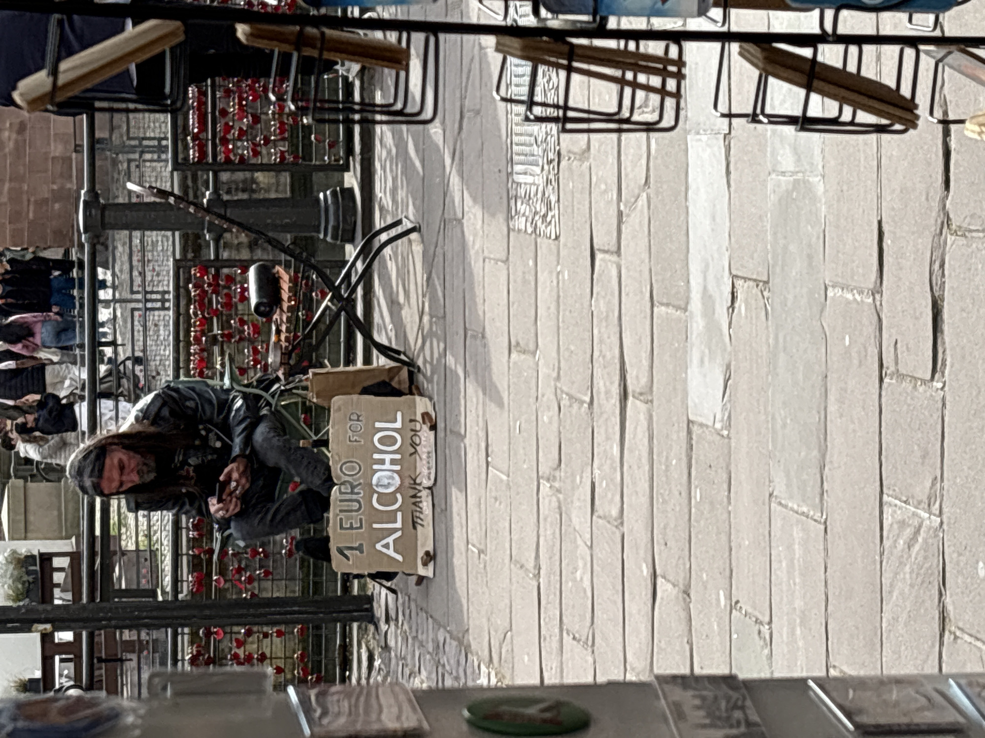 The image shows a man with long hair and a beard, wearing a dark jacket and pants, squatting on a brick-paved street. He is holding a mobile phone in his hands. Next to him, there's a sign that reads "1 EURO FOR ALCOHOL, THANK YOU". The background suggests a busy, public area with metal structures and other people in the periphery. The scene is likely in Strasbourg, France, judging by the architectural details and the typical cobblestone pavement found in many European city centers. The general impression is of a street performer or beggar asking for money.