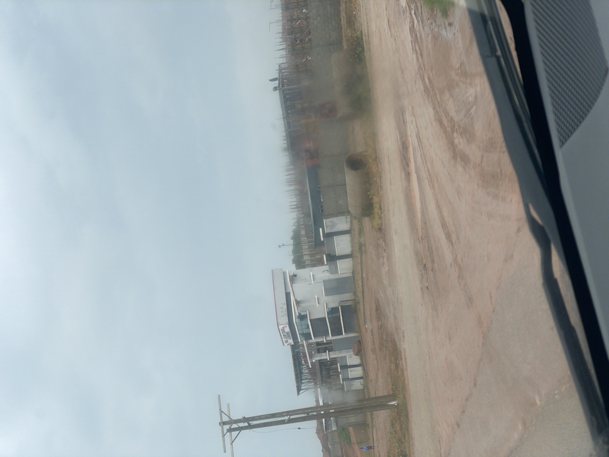 The image depicts a construction site in Kurunduma, Nigeria, under a cloudy sky. In the foreground, a dirt road with tire tracks is visible, and the edge of a vehicle's windshield and dashboard is present. A utility pole stands to the left, with wires extending from it. Behind the pole, a partially constructed building with a white facade and a sign that reads "PHIL LUXURY ESTATE AKWOKORO" is situated. Further back, more buildings are under construction, with visible concrete pillars and rebar. Several workers are present on the upper levels of these structures. In the middle ground, two large concrete pipes lie on the ground, and dust is being kicked up, likely from a vehicle or activity not fully captured in the frame. A single person is walking on the left side of the road. The environment appears to be an active development area in a semi-urban setting.