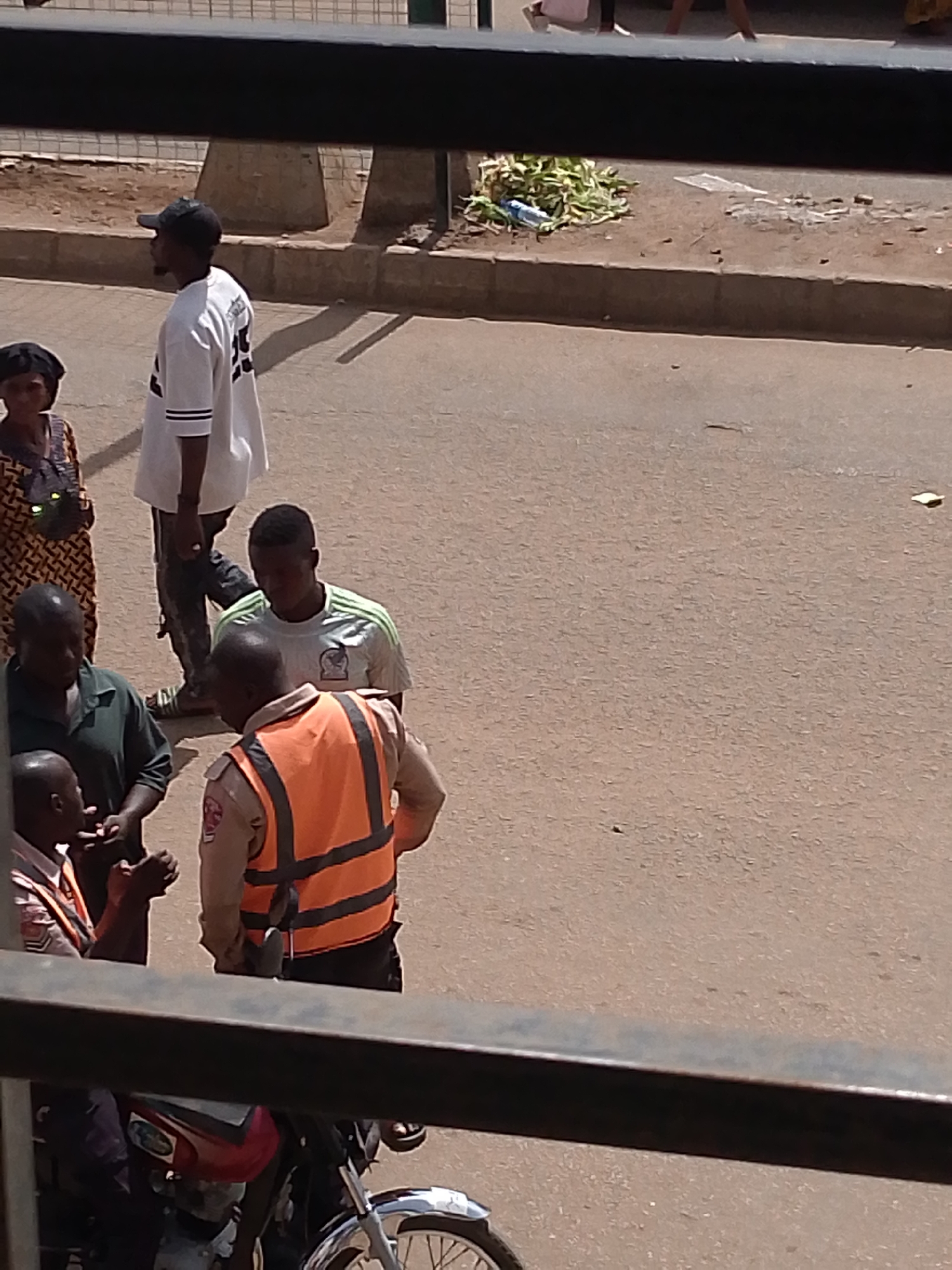 In Bukuru, Nigeria, a group of individuals is gathered on a paved street. In the foreground, a person wearing an orange vest over a tan uniform is speaking with two other men. One of these men is wearing a dark green shirt, and the other is partially obscured but appears to be wearing a light-colored shirt. Behind them, another man in a light green shirt is looking down. Further back, a man in a white t-shirt and a black baseball cap is walking away from the group. To the left, a woman in a patterned dress and head covering is standing. The background shows a dirt area with some vegetation and a curb. A motorcycle is visible in the lower left corner.