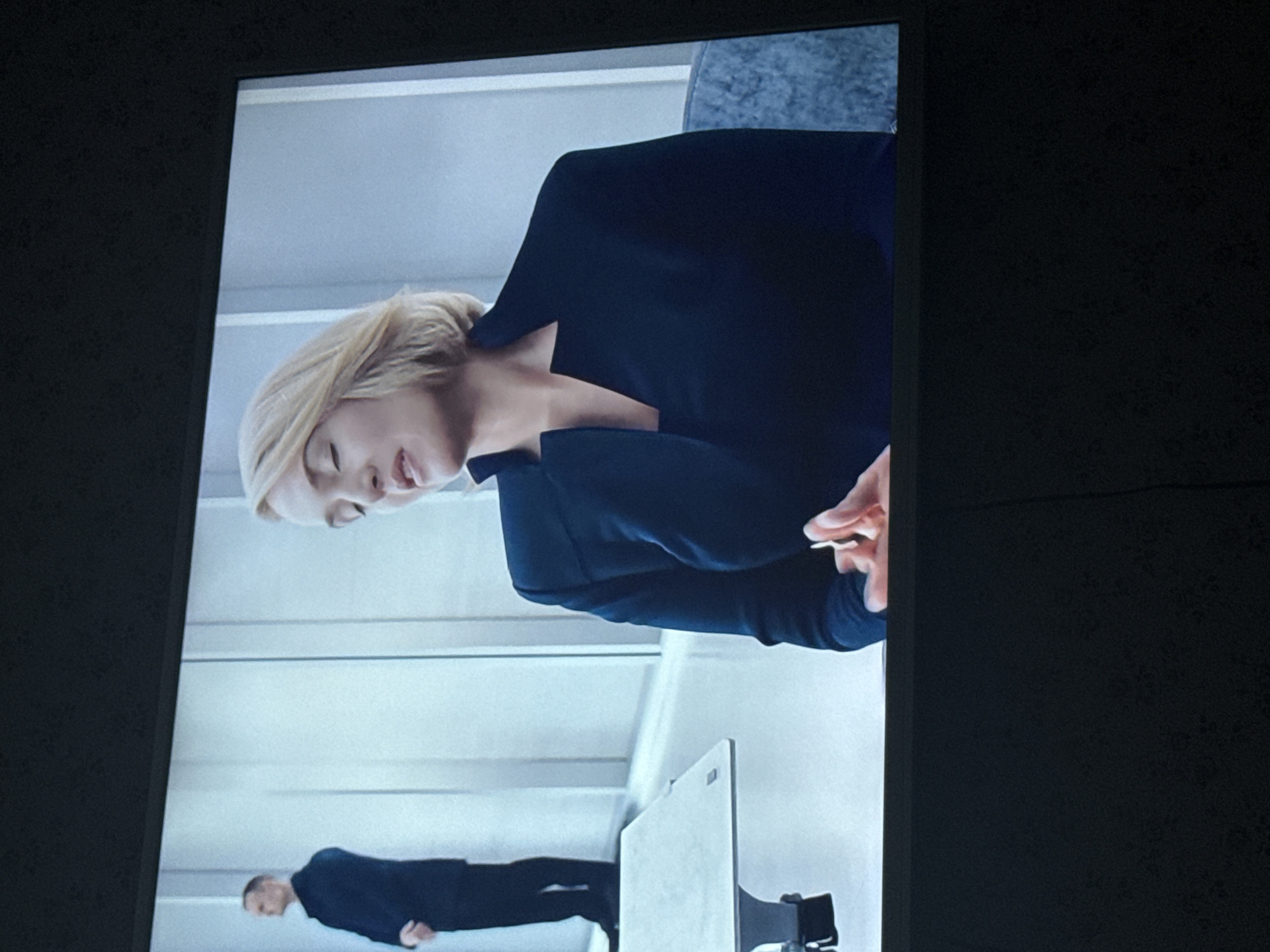 The image shows a screen displaying a scene with two people in a modern, white interior. At the top, a woman with short blonde hair, wearing a dark suit, is looking down with her eyes partially closed. Her hand is visible, holding something small and light-colored. Below and further away, a man in a dark suit is standing. In the foreground is a light-colored object resembling a modern desk or screen with a stand. The setting appears to be an office or public space with clean lines and neutral colors. The location is not identifiable from the image.