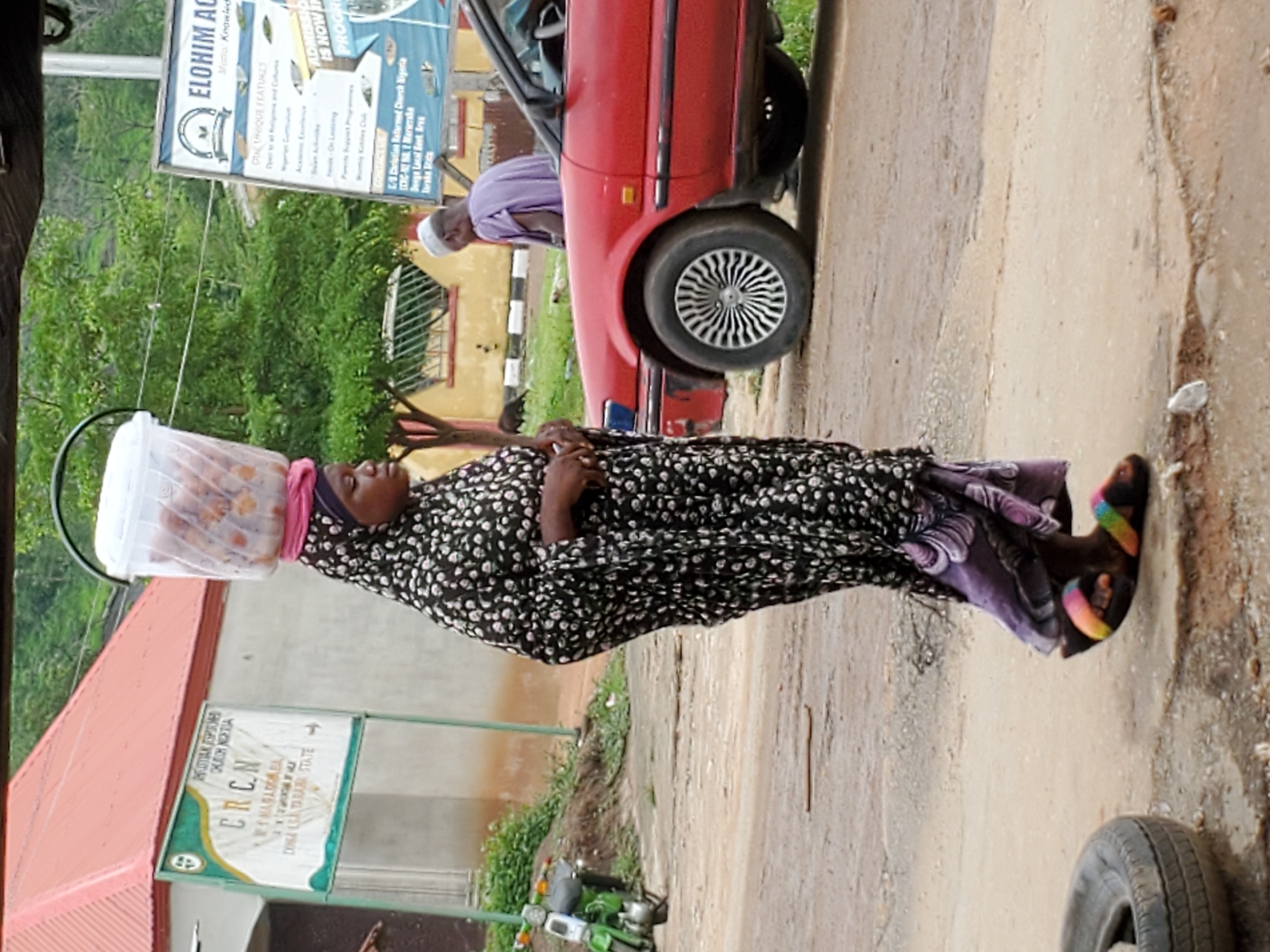 The image captures a street scene in Mararaba, Nigeria, during the daytime. The main subject is a woman walking on the right side of an unpaved road, expertly balancing a transparent plastic container on her head. The container appears to hold items, possibly food like bread or pastries. She wears a black and white patterned long dress, a dark headscarf, and a pink fabric beneath the container, along with colorful sandals. Her expression is neutral as she looks forward.
Behind a parked red car on the left, another person, likely a man in a light purple garment and white cap, is partially visible, appearing to attend to the car's interior. The red car has its driver's side door open. Green foliage and trees are prominent in the background, suggesting a natural environment alongside the road. A building with a reddish roof is partially seen on the far left. A green motorcycle or scooter is also visible on the ground.
The road surface is unpaved or made of worn concrete, showing cracks and dirt. A discarded tire lies near the bottom right.
Two signs are visible:
1. A blue and white sign, partly obscured, advertises "ELOHIM AC" with the motto "Knowledge". It lists "UNIQUE FEATURES" and mentions "ADMISSION IS NOW OPEN". Crucially, it identifies "Christian Reformed Church Nigeria (CRC-N) No. 1 Mararaba, Denga Local Govt. Area, Taraba State," confirming the location.
2. A green and white sign, also for the "CHRISTIAN REFORMED CHURCH NIGERIA (CRCN)", indicates "NO 1 MARARABA OLD DANKO LGA TARABA STATE".