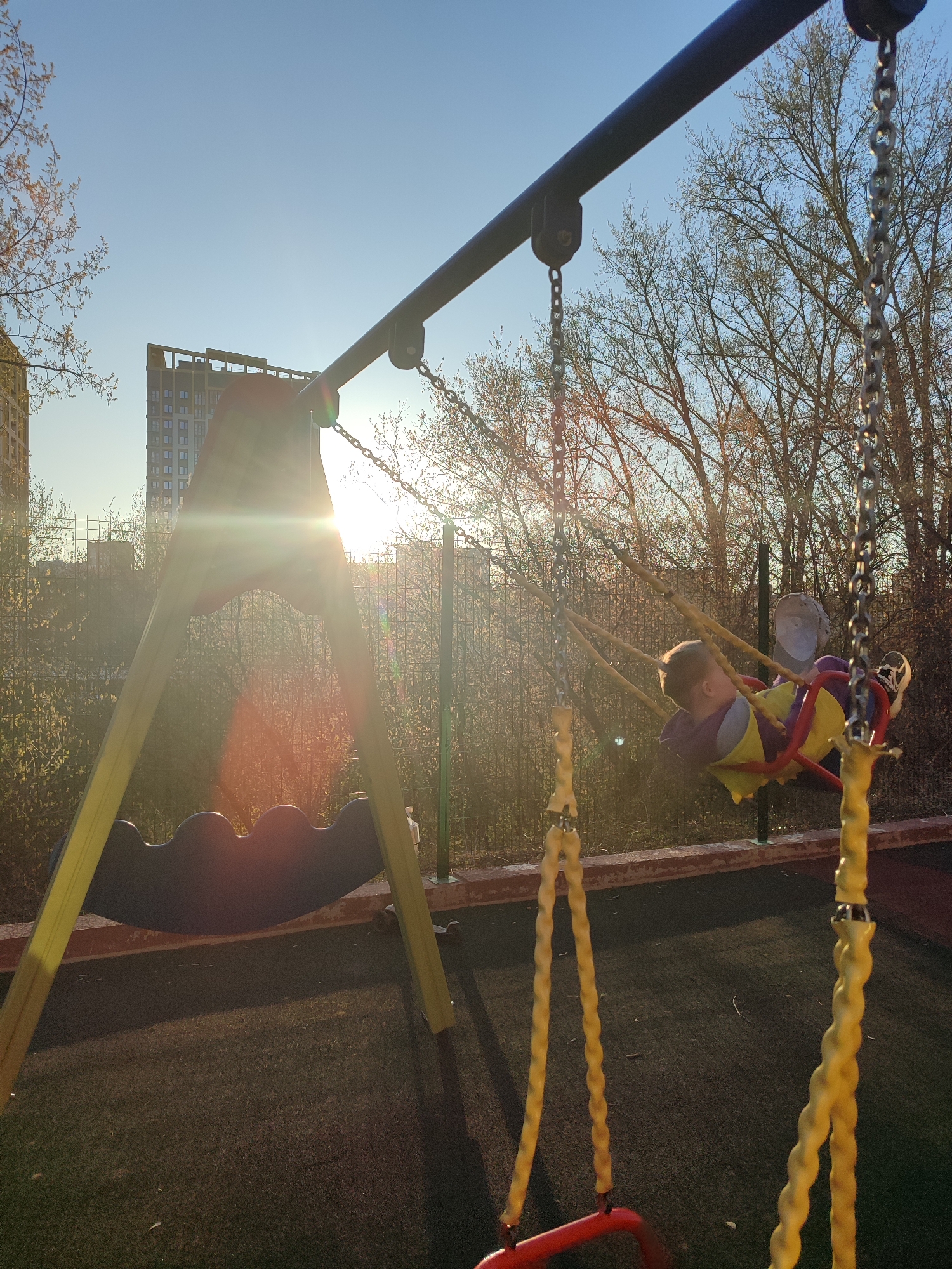 A sunny outdoor playground scene in Izhevsk, Russia, features a child on a yellow and red swing. The swing set is positioned in front of a fence with green vegetation behind it, and bare trees are visible beyond the fence. In the background is a multi-story building under a blue sky, partially obscured by sunlight. A second piece of playground equipment sits to the left of the swing. The playground has a dark ground surface, likely a safety material.