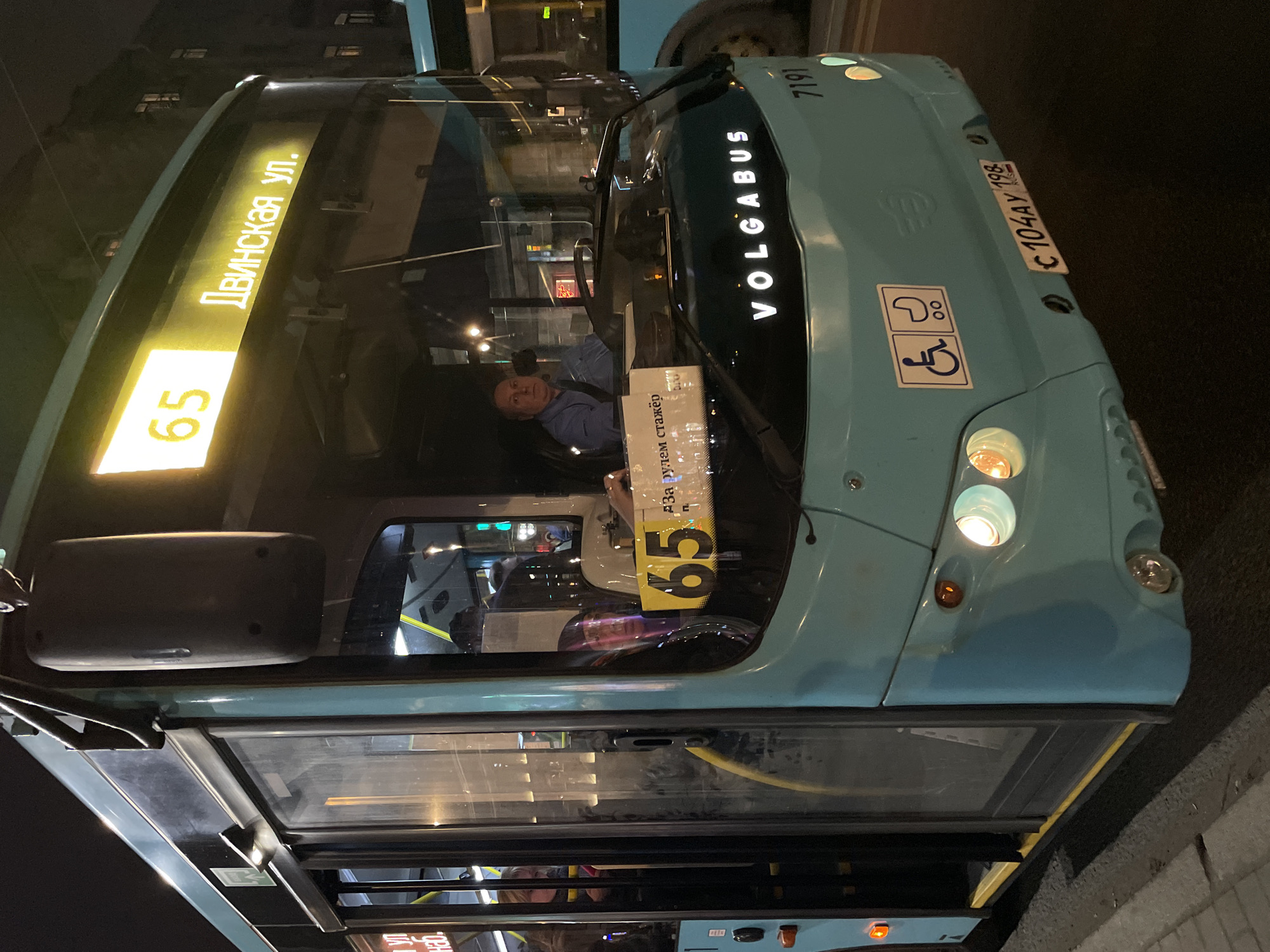 The image shows a blue Volgabus bus, route number 65, with the destination sign reading "Двинская ул." (Dvinskaya Street) in Saint Petersburg, Russia. The bus is displaying the license plate "C 104 AY 198". The bus is marked with wheelchair access symbols. The bus driver is visible inside the vehicle. A sign in Russian, visible on the windshield, indicates that the driver is a trainee. The image was taken at night.