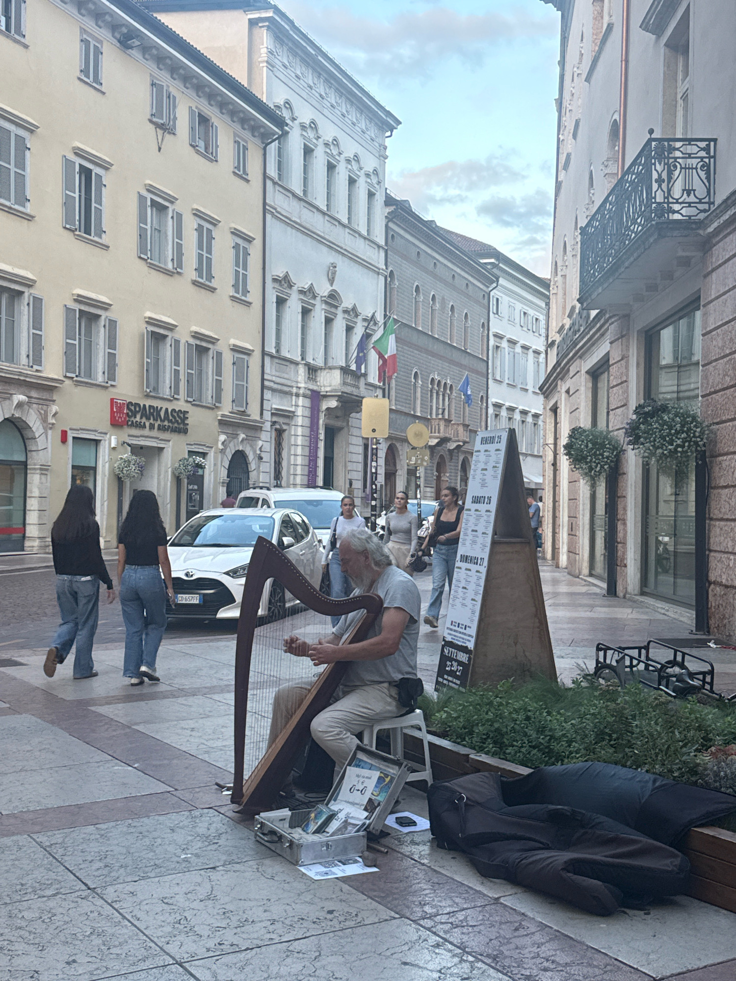 An elderly man with gray hair and a beard is playing a harp on a cobblestone street in Trento, Italy. He is seated on a small white stool, wearing a light-colored short-sleeved shirt and light-colored pants. His right hand is plucking the harp strings. In front of him is an open silver case containing some CDs and money. A black fabric bag is resting on the ground to the right of the stool.
Behind the harpist, two young women with dark hair are walking away from the camera. The woman on the left is wearing a dark long-sleeved shirt and blue jeans. The woman on the right is wearing a black t-shirt and blue jeans.
Further back, a white car is parked on the street. The building on the left has a sign that reads "SPARKASSE CASSA DI RISPARMIO". On the right side of the street, there is a pyramid-shaped sign with text in Italian. The street is lined with buildings with multiple stories and windows. The sky above is overcast with some clouds. The scene appears to be a street in a European city, likely a pedestrianized area.