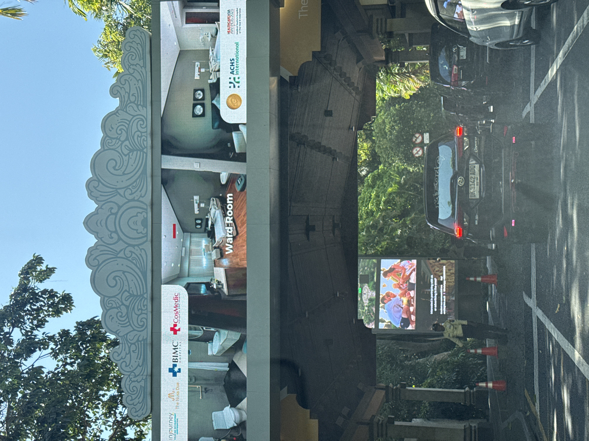 In Kuta Selatan, Indonesia, a traffic scene unfolds under a bright, clear sky, with lush green trees framing the top portion of the image. A large digital billboard arching over the road displays advertising content. The top section of the billboard shows a "Ward Room" advertisement for BIMC CosMedic Hospital, featuring images of modern hospital rooms and bathrooms, along with logos for "injourney" and "ACHS International." Below this, another billboard displays an advertisement for "The Nusa Dua," featuring a family enjoying an outdoor activity and text encouraging partnership and advertising. In the foreground, a traffic controller in a yellow uniform stands near orange traffic cones, directing vehicles. Several cars are stopped or moving slowly in traffic, including a black SUV with a license plate reading "DK 1142 ADQ," a black Innova, and a white vehicle. The road surface is asphalt with white lane markings. The overall scene depicts a typical traffic flow in a tropical, commercial area.