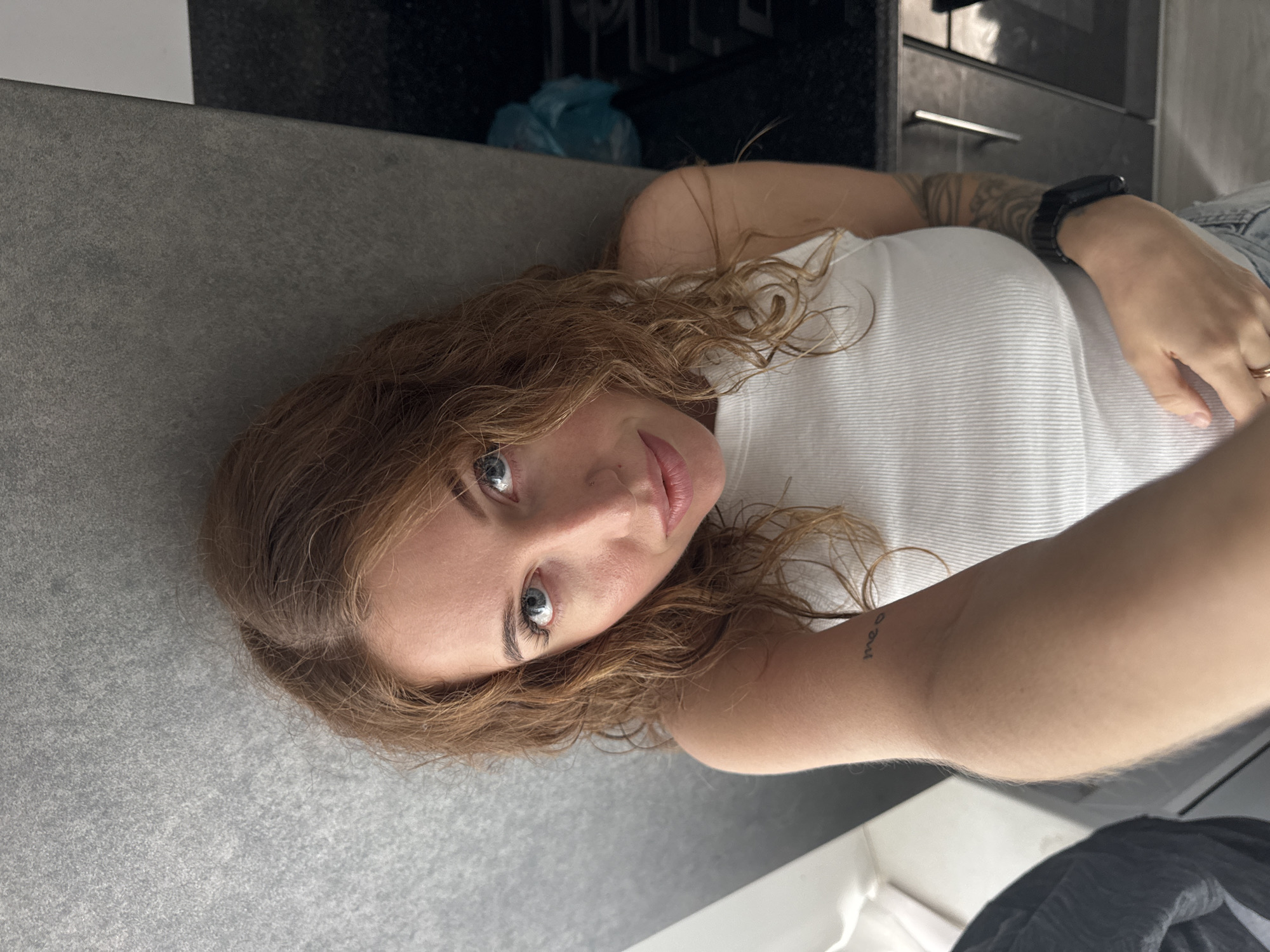 The image shows a young woman taking a selfie in an indoor setting, likely a kitchen, in Chaikovsky, Russia. She is positioned horizontally, looking directly into the camera with a calm expression and a slight smile. Her medium-length, wavy, light brown hair frames her face, highlighting her blue eyes. She is wearing a white ribbed tank top and light-colored denim pants.
Visible accessories include a dark-colored smartwatch on her left wrist and a ring on her right ring finger. She has two tattoos: a small text tattoo on her left forearm reads "meow," and a larger, abstract or tribal pattern tattoo is visible on her upper left arm/shoulder area.
The woman is lying or leaning on a gray, textured countertop surface that dominates the foreground and left side of the image. In the background, a dark kitchen area is visible, featuring what appears to be an oven or cabinet with handles, and a light blue plastic bag. The overall lighting suggests it is daytime.