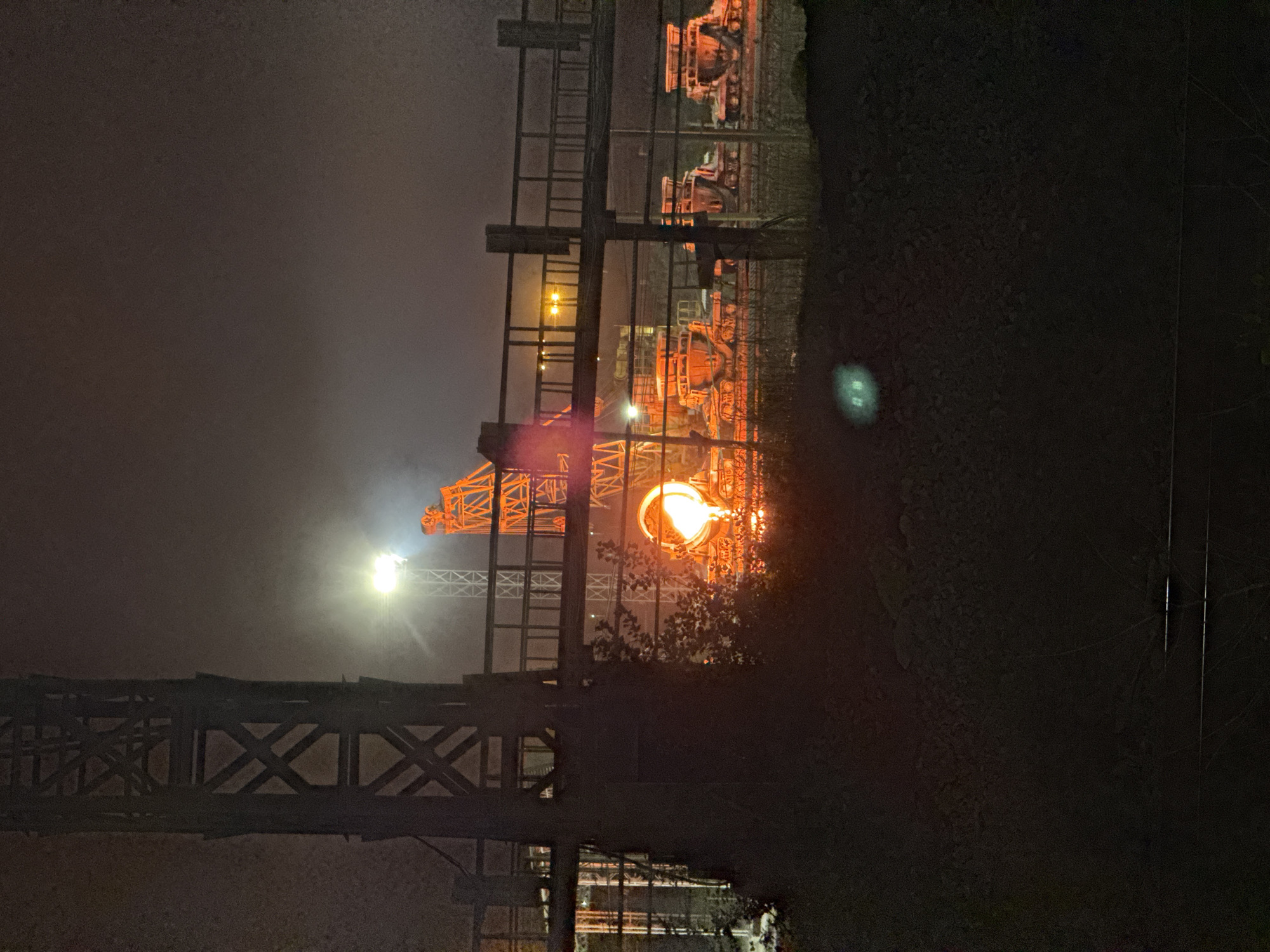An industrial scene at night in Magnitogorsk, Russia, featuring a large structure that appears to be a blast furnace, with molten metal glowing orange. A large metal tower is visible on the left side of the image. There are also several industrial vehicles or carts that appear to be orange. A walkway or bridge structure runs horizontally across the middle of the image. The overall environment is dark and hazy, with some bright lights illuminating the scene. The ground in the foreground is covered with dark material, possibly coal or slag.