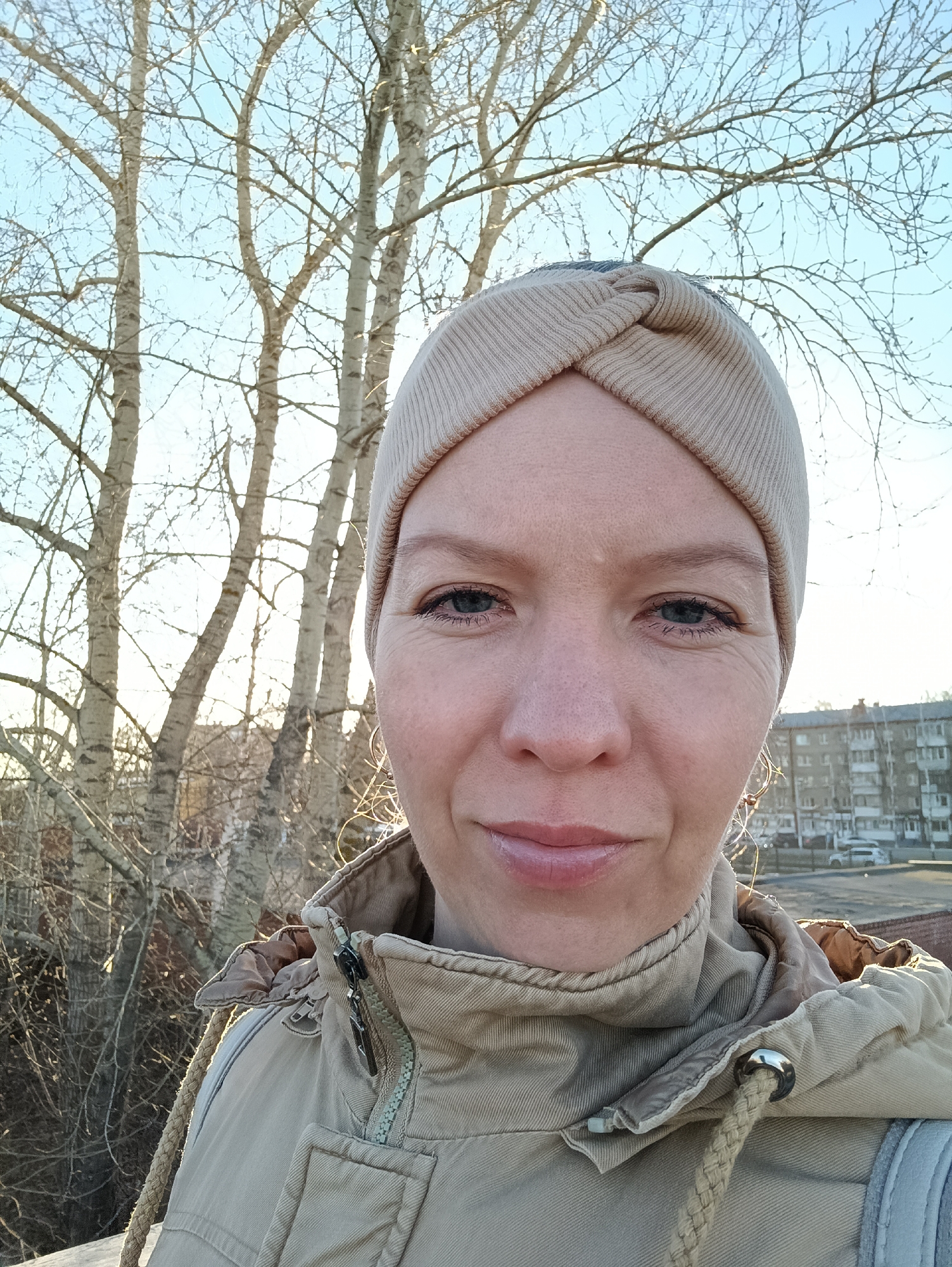 A light-skinned woman wearing a beige head wrap and a beige hooded jacket takes a selfie in an outdoor setting. The background includes bare trees with white trunks and a building with multiple windows and parked cars, typical of urban residential areas. The location is likely in the city of Chaikovsky, Russia. The woman's expression is neutral, and the lighting suggests it is daytime. The jacket is zippered, with drawstrings and a hood visible.