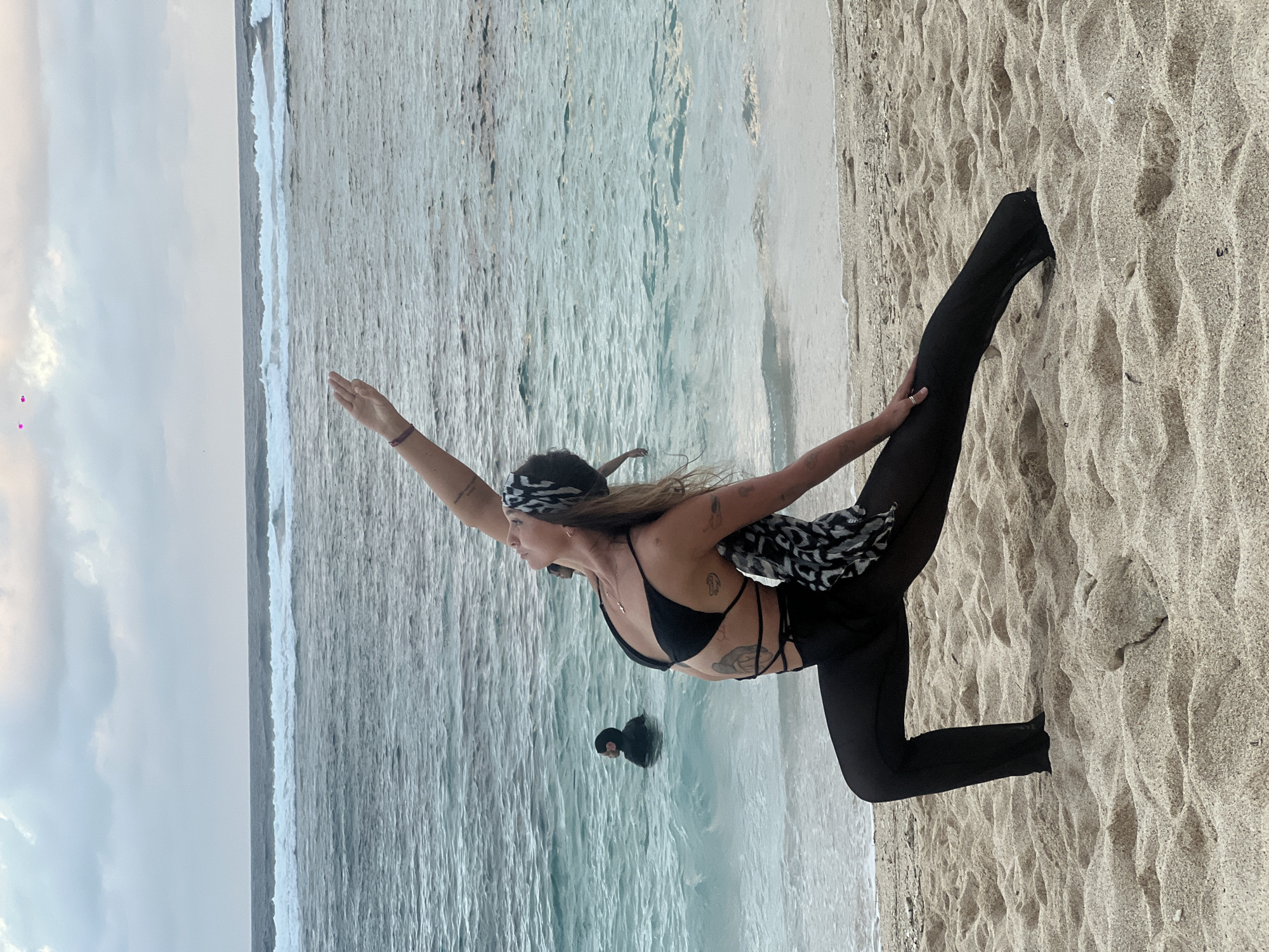 A woman in a black bikini top, black leggings, and a patterned headscarf performs a yoga pose on the beach in Kuta Selatan, Indonesia. Her left leg is straight and grounded in the sand, while her right leg is extended upwards. Her left arm reaches forward and her right arm extends towards her raised leg. There is a person in the water nearby. The scene includes the ocean, waves, sand, and a cloudy sky.