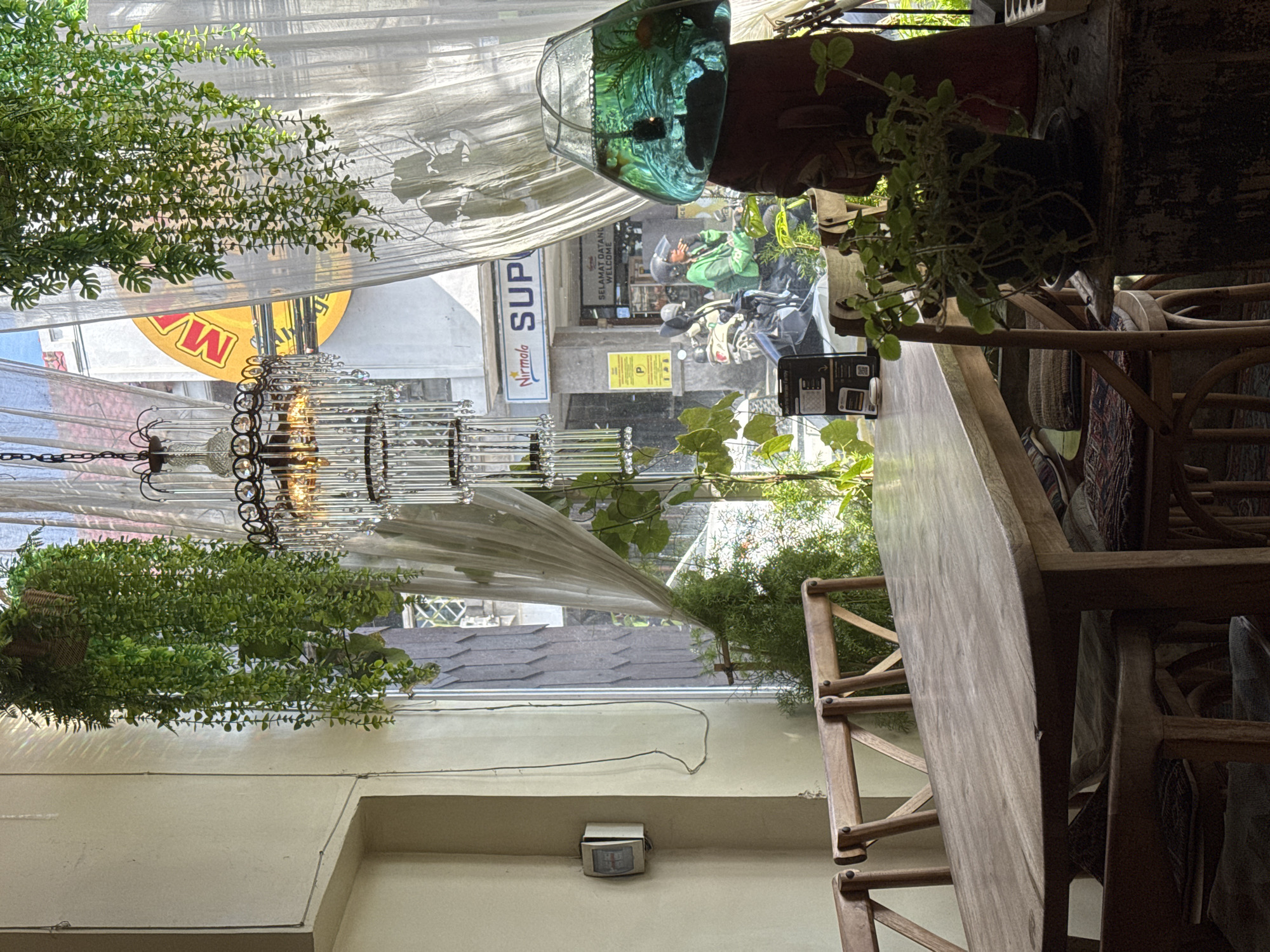 The image depicts the interior of a restaurant or cafe, possibly in an urban setting. The restaurant's exterior features a sign for "Nirmala SUP," indicating a possible commercial establishment. An individual wearing a green uniform and a helmet, likely a delivery person, is visible outside on a motorbike. Inside, there is a long wooden table with chairs, suggesting a dining area. Hanging plants and a chandelier add to the decor, and sheer curtains adorn the windows, creating a soft, diffused light. The location is indicated as "Unknown, Unknown."