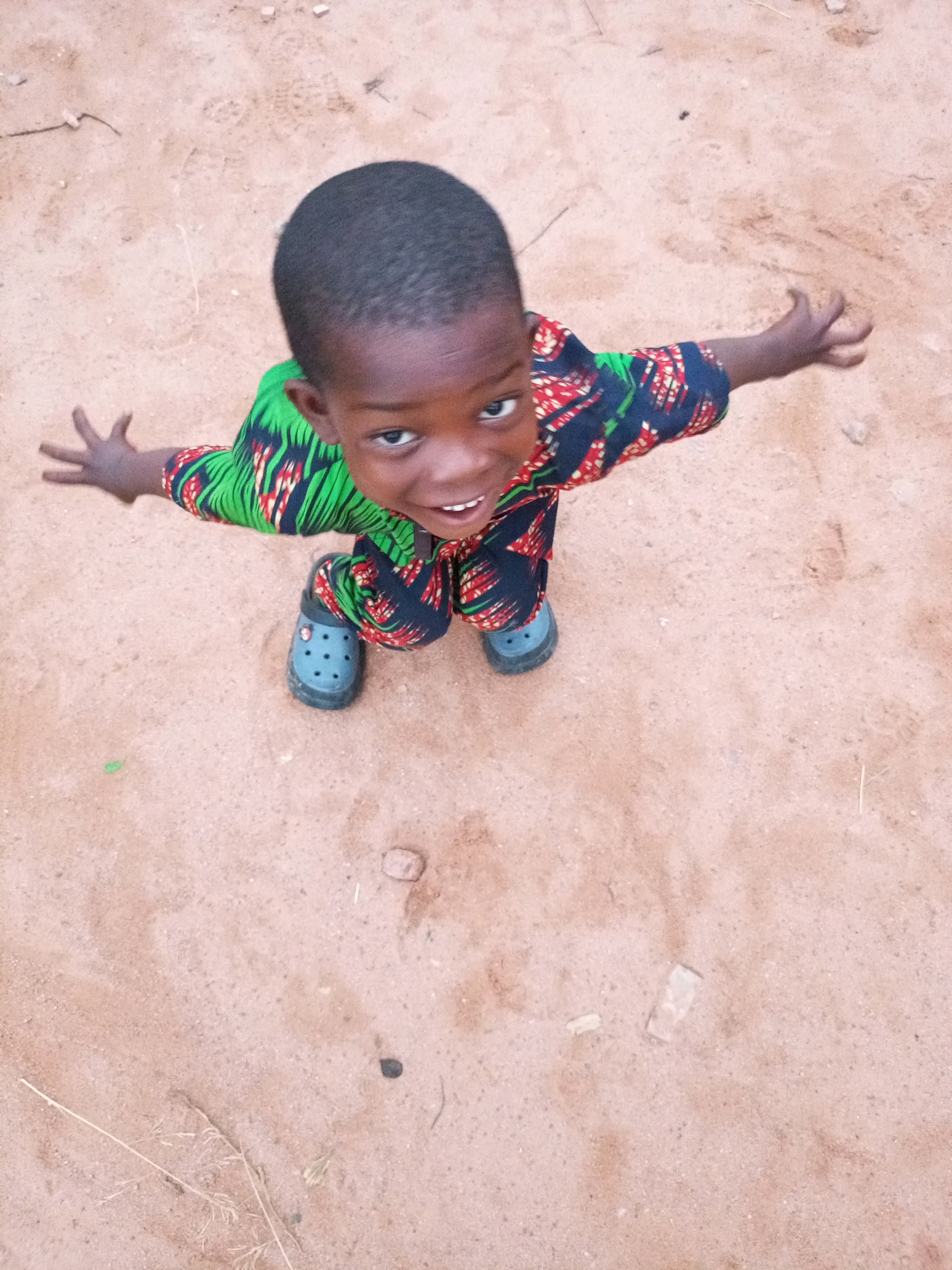 A young child, appearing to be of African descent, is positioned in the center of the frame, looking upwards towards the camera. The child has dark skin and short, dark hair. They are wearing a garment with a vibrant, abstract pattern of green, red, and black. The child's hands are outstretched, palms facing upwards, in a gesture of reaching or offering. On their feet are dark grey clogs. The child is standing on a surface that appears to be dry, sandy ground, with scattered small stones and dried vegetation. The lighting suggests it is daytime, with the sun casting a gentle light. The background is an out-of-focus expanse of the same sandy ground. The scene has been captured from a high-angle perspective. The overall setting suggests an outdoor environment, possibly in a rural or less developed urban area, which aligns with the specified location of Tempure, Nigeria.