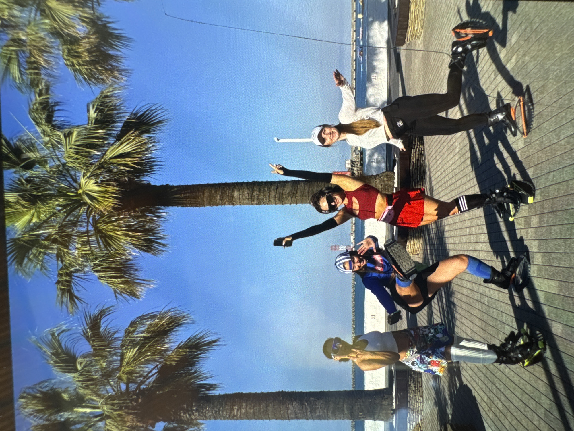 Four women are posing on a wooden boardwalk in Sochi, Russia. They are all wearing athletic clothing and specialized footwear, possibly for an exercise activity. The woman on the far left is wearing a white tank top and a colorful skirt, with white knee-high socks. The second woman from the left is wearing a blue jacket with a striped hat and blue socks. The third woman from the left is wearing a red sports bra and a red skirt with black and white striped socks. The woman on the far right is wearing a white long-sleeved shirt and black pants. Two of the women are holding their phones up, possibly taking pictures or videos. Palm trees are visible in the background, along with a beach and the sea. The sky is clear and blue. The overall scene suggests a sunny day and an outdoor recreational activity.