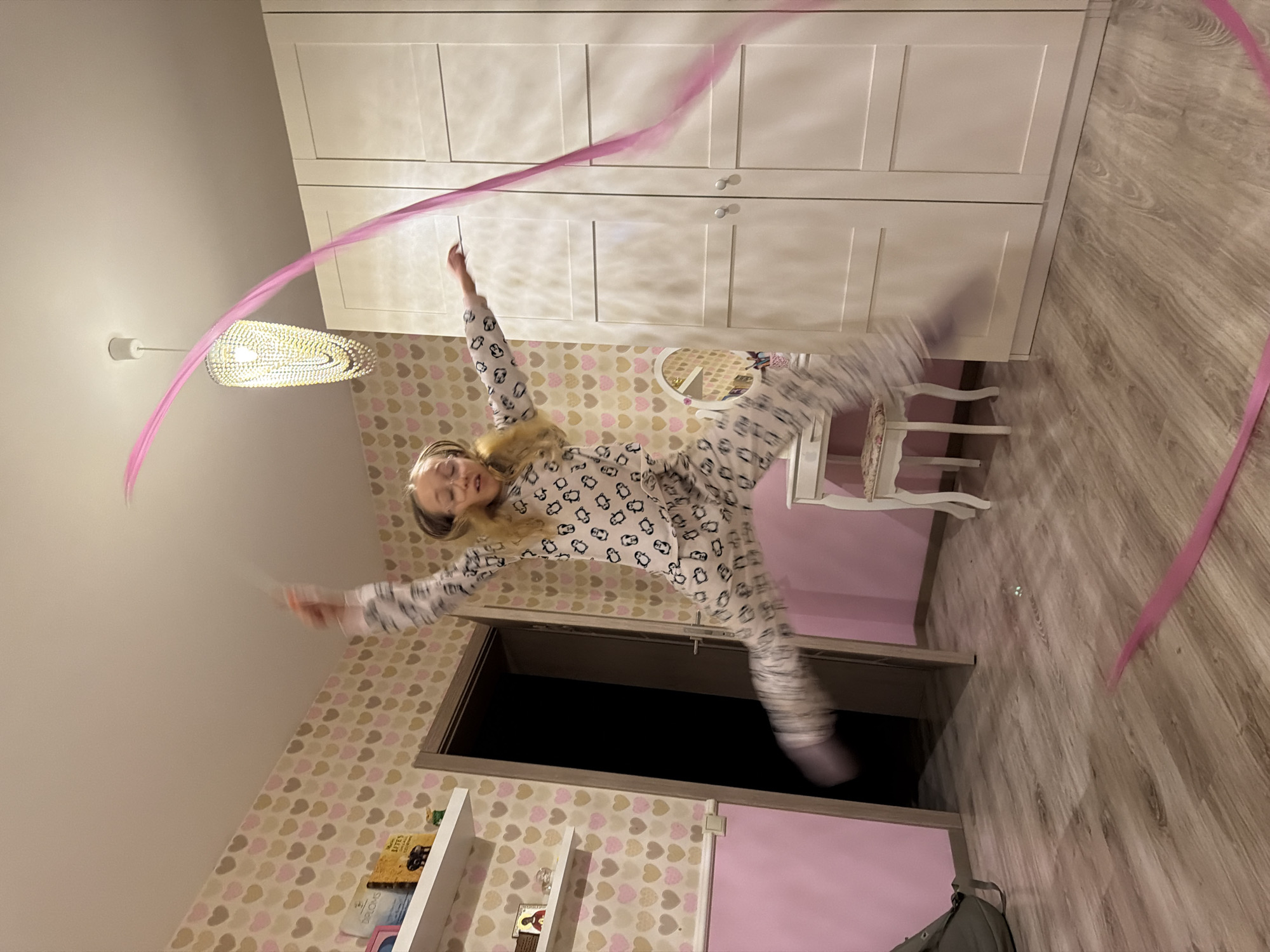 A young girl in penguin-patterned pajamas leaps with a pink ribbon in what appears to be a bedroom. The room is decorated with heart-patterned wallpaper and features a white wardrobe, a small white chair, and a bookshelf. The lighting is warm, suggesting evening. The location is most likely a residential setting in Dreiliņi, Latvia. The girl's arms are outstretched, holding the ribbon, which creates a sweeping arc in the frame.