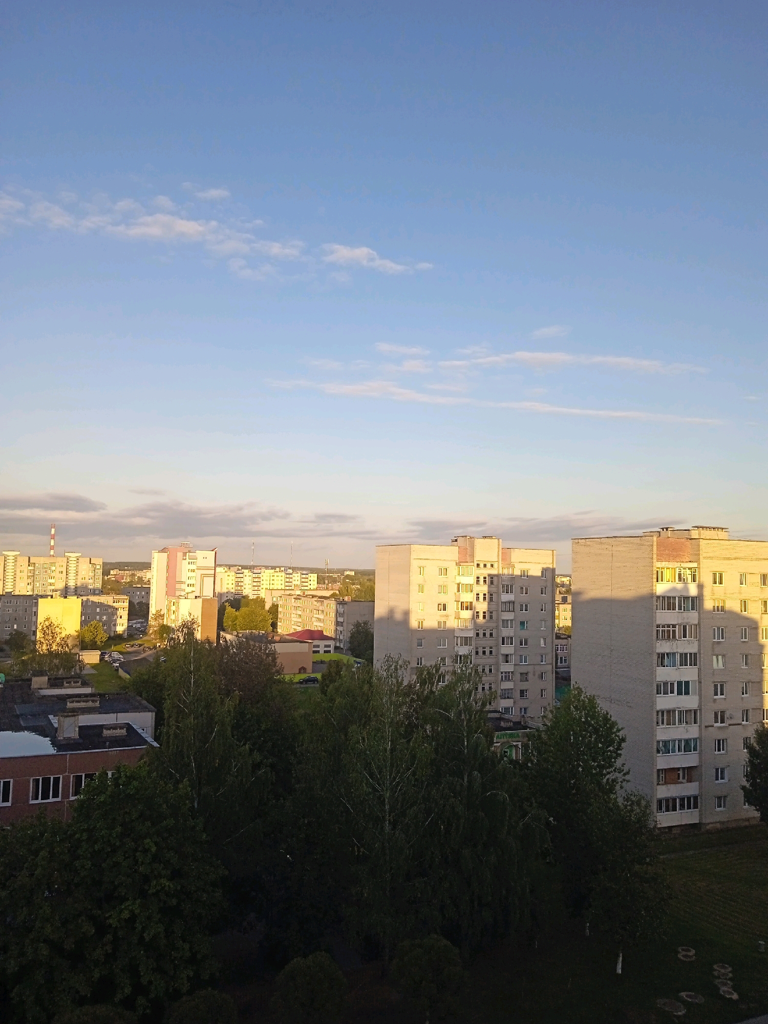 The image shows a cityscape in Lida, Belarus, likely captured during late afternoon or early evening due to the long shadows and warm lighting. The scene is dominated by multi-story apartment buildings of varying architectural styles, characteristic of Eastern European urban planning. Some buildings are lighter in color, possibly concrete or painted, while others appear to be brick. They are primarily residential, with numerous windows visible.
In the foreground, a dense canopy of mature trees, predominantly deciduous with some birch trees, creates a dark, leafy barrier. These trees are interspersed with smaller buildings and what appear to be parking areas or courtyards. The ground in the immediate foreground, particularly on the right side, is covered in grass with circular markings, possibly a children's play area or sports court.
Further back, more apartment blocks are visible, stretching into the distance. One prominent tall building in the middle ground has a pink and white facade. In the background, a tall industrial chimney with red and white markings stands out against the sky. The sky itself is a clear, light blue with scattered, wispy white clouds, and the setting sun casts a warm glow on the buildings and trees, creating strong contrasts between light and shadow. No people are visible in the frame. The overall impression is of a typical residential district within a Belarusian city.