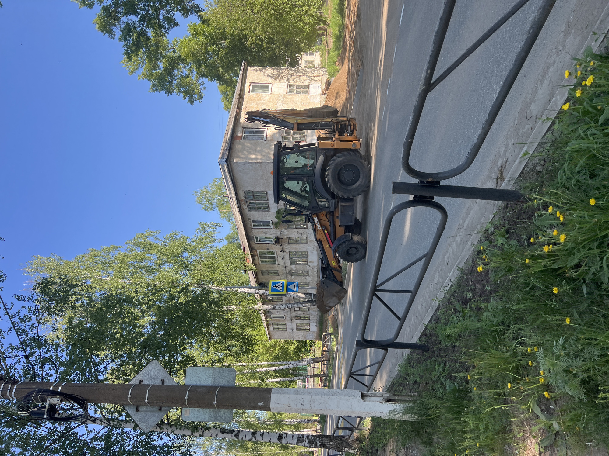 The image depicts a backhoe parked on the side of a road next to a building in Chaikovsky, Russia. The building is a low-rise apartment building with multiple windows. A pedestrian crossing sign is visible near the building. There are trees lining the street. A metal fence runs along the sidewalk. Dandelions grow in the grass next to the sidewalk. An electrical pole is located on the left side of the image.