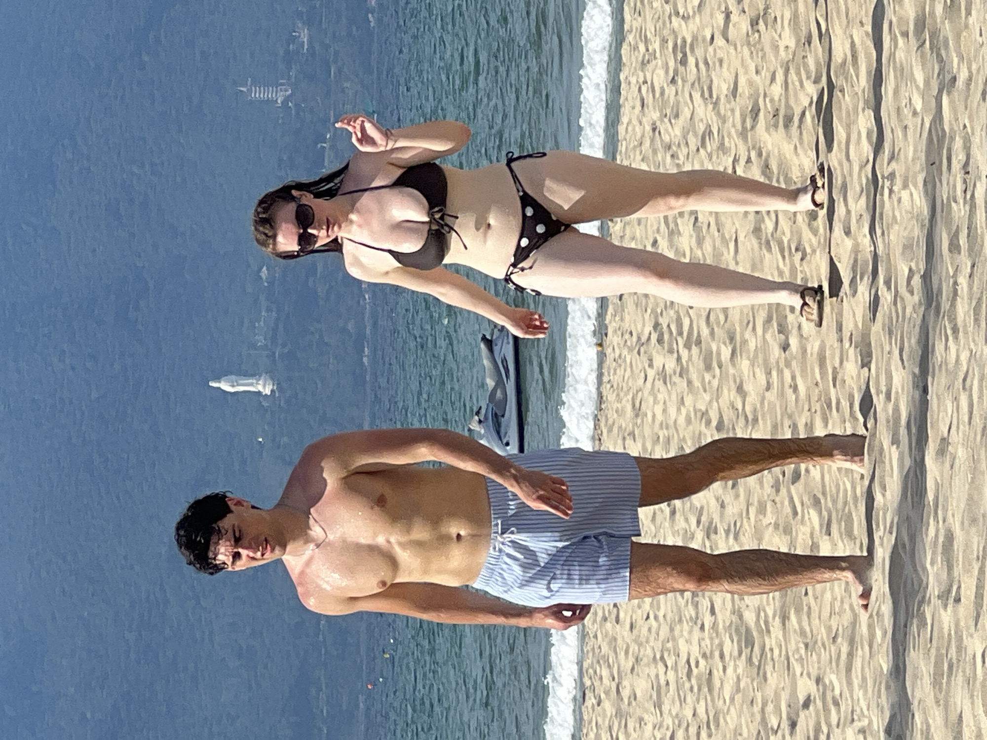 Two individuals are walking on a sandy beach. On the left, a man with dark hair, wearing blue and white striped swim shorts, stands looking downwards. He is bare-chested and appears to have water droplets on his skin. On the right, a woman with dark hair, wearing a black polka dot bikini and sunglasses, walks with her right hand raised slightly. Behind them, the ocean is visible with waves breaking near the shore. In the background, a jet ski is partially submerged in the water, and further in the distance, a white statue of a figure can be seen on a hillside, possibly the Lady Buddha statue at the Linh Ung Pagoda complex on Sơn Trà Peninsula in Đà Nẵng, Vietnam. The sky is a clear blue.