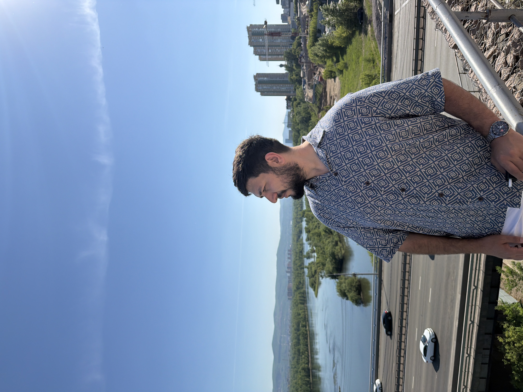 The image depicts an outdoor, elevated daytime scene overlooking a significant river, urban infrastructure, and distant cityscape. A man is positioned in the right foreground, captured from the mid-chest upwards, with his head turned to the left, observing the panoramic view. He has dark hair and a beard and wears a short-sleeved, collared shirt with a repetitive geometric pattern in blue and off-white. A watch is visible on his left wrist.
In the middle ground, below the man, a multi-lane highway or bridge extends across the frame. Two white cars and one dark car are visible on the road, with barriers separating the lanes. Below the highway, a wide river, identified as the Yenisei River in Krasnoyarsk, Russia, dominates the landscape. Several small, densely vegetated islands are scattered within the river. The riverbanks are lined with lush green trees, extending into forested areas. On the distant left bank, a range of green mountains or hills outlines the horizon, typical of the landscape surrounding Krasnoyarsk.
To the right of the river, on the closest bank, a construction site is visible, featuring at least two tall cranes and excavated ground. Behind the construction, a cluster of modern high-rise residential buildings, characterized by their uniform, tall structures, stands prominent against the sky. A small, dark dome-shaped structure, possibly a church, is discernible between two of these buildings. The sky above is clear and bright blue with some faint, wispy cirrus clouds or contrails stretching horizontally across the upper portion of the frame. The scene is brightly lit by direct sunlight.