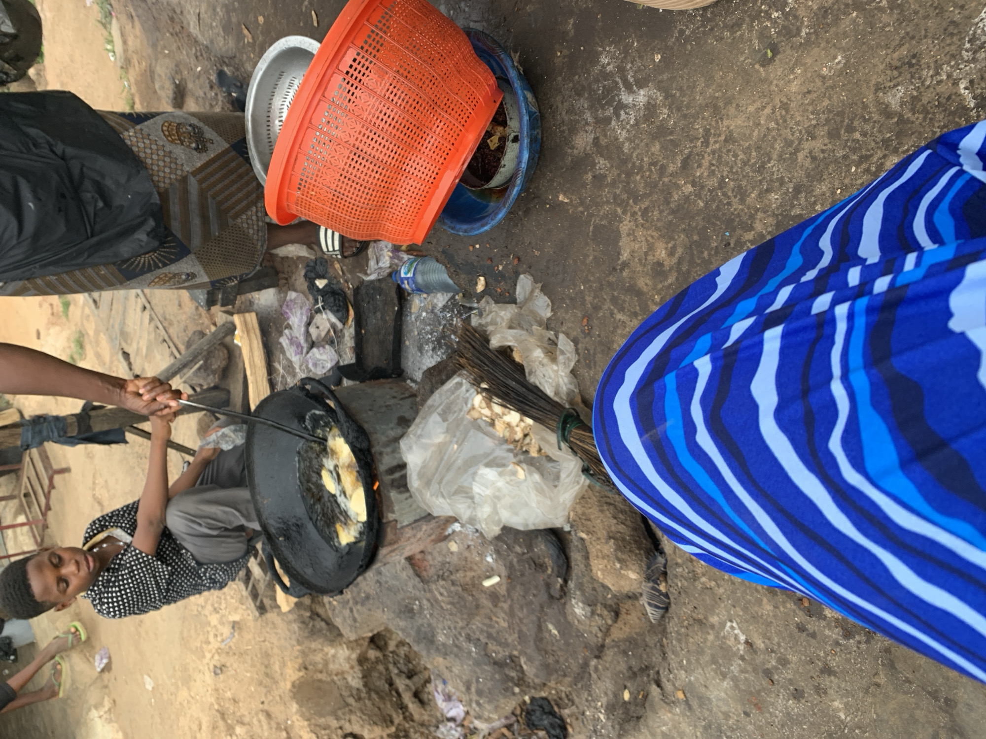 A young individual is positioned next to a cooking setup, in an outdoor environment near New Stadium, Nigeria. The individual is squatting or sitting low and operating a large, blackened pan on a crude stone or brick structure. The pan contains what appears to be food being cooked, potentially fried. A pair of hands is visible above the pan, likely guiding or assisting with the cooking process using a long tool. Next to the cooking setup, there is an assortment of items: an orange basket, a silver colander, a blue container, a broom and various plastic bags, some of which appear to contain food items or ingredients. The ground is uneven and appears to be a dirt or concrete surface. A patterned fabric or covering is draped nearby, possibly functioning as a workstation or for storage.