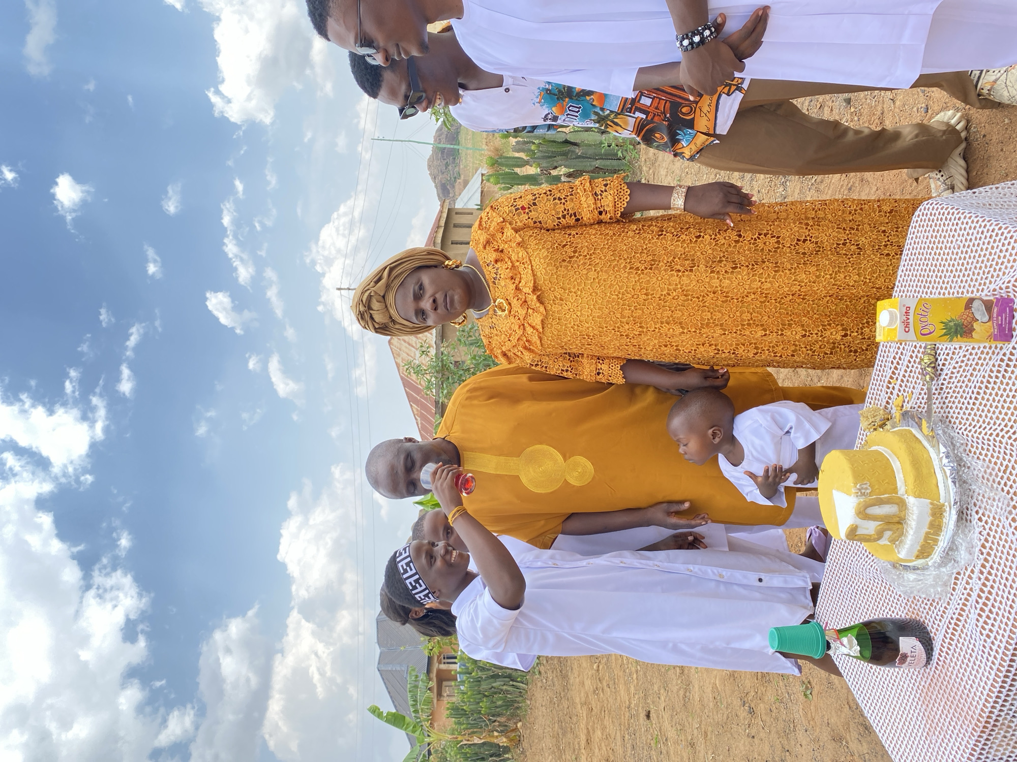 The image captures a family celebration outdoors in Kuru Karama, Nigeria, under a partly cloudy sky. A group of people, including adults and a child, are gathered around a table. The table has a white netting cloth covering it, and on it, there's a cake decorated in yellow, a Chivita juice box, and an uncorked bottle of what appears to be sparkling wine with a green plastic cup used as a stopper. Individuals are dressed in colorful, traditional attire, with some wearing yellow garments and others in white. One person is holding a cup containing a red liquid. The setting seems to be a residential area with houses and greenery in the background.
