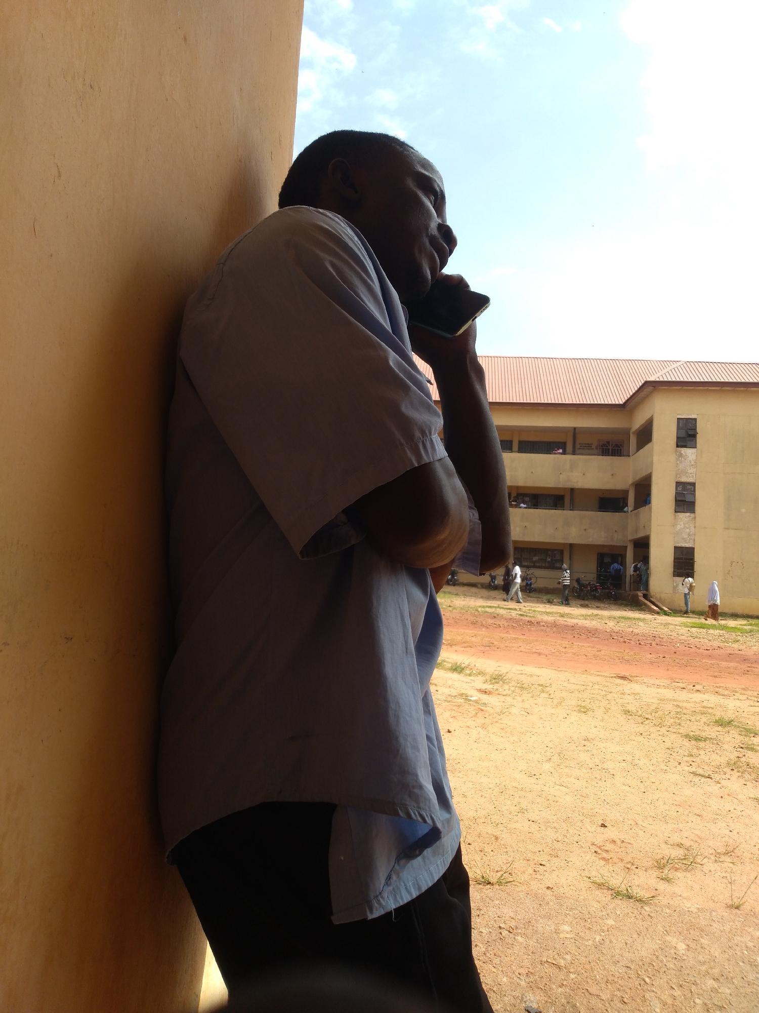An outdoor, low-angle, medium shot shows a dark-skinned man in a light blue, collared shirt and black pants, leaning against a textured, ochre-colored wall. He is facing away from the camera, looking to his right, and holding a dark-colored mobile phone to his right ear with his right hand. The background reveals a dusty, unpaved courtyard in front of a multi-story, yellow building with a red roof. The building has numerous windows, and several people, some appearing to be students, are visible on the ground and on balconies. The sky above is bright and partly cloudy. This scene is likely located in a school or institutional setting in Jos, Nigeria, given the architecture and the presence of people dressed in a manner consistent with the region.
