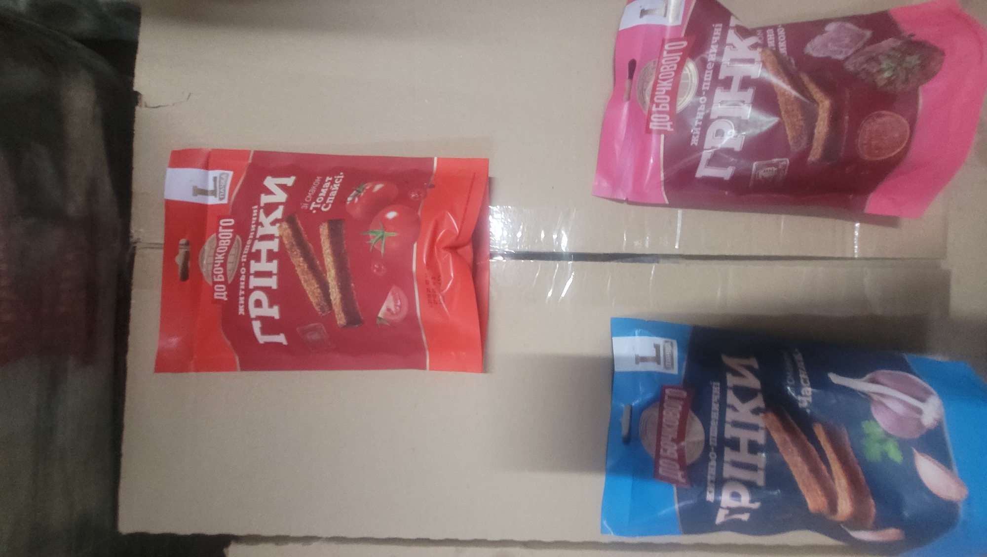 This is an image of four bags of croutons, or "grinki" in Ukrainian, placed on a cardboard surface. The bags are oriented in different directions and are primarily red, pink, and blue.
The red bag in the upper left features text in Ukrainian, "ДО БОЧКОВОГО ЖИТНЬО-ПШЕНИЧНІ ГРІНКИ", which translates to "For Barrel, Rye-Wheat Croutons". It also shows images of tomatoes and slices of tomato. The pink bag, positioned above and to the right, has similar text and imagery, including what appears to be dried meat or jerky and herbs. The blue bag in the lower right corner displays the text "ДО БОЧКОВОГО ЖИТНЬО-ПШЕНИЧНІ ГРІНКИ" with the flavor indicated as "Часник" (Garlic), and features images of garlic cloves and parsley. Another bag, partially visible in the top right corner, is pink and also displays Ukrainian text.
The croutons themselves are visible in some of the bags, appearing as toasted bread slices. The overall scene suggests a product display or inventory. The lighting is somewhat dim, and the focus is on the bags of croutons. The cardboard surface appears to be a plain, neutral background. There are no people or animals present in the image.