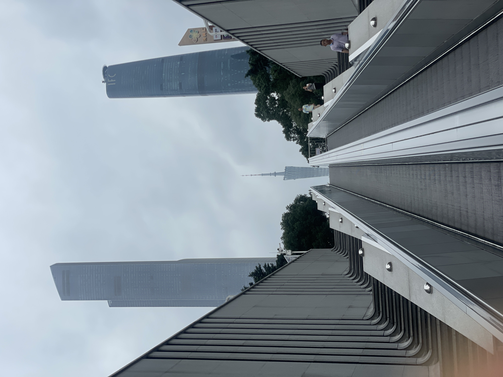 The image shows a view in the Tianhe District of Guangzhou, China. The perspective is upward from a structure with a series of upward-sloping walkways with metal railings and integrated lighting. In the background, several skyscrapers are visible, including one labeled "IFC." A tall, thin spire is located between the buildings. A few people are visible on the walkways. The sky is overcast.