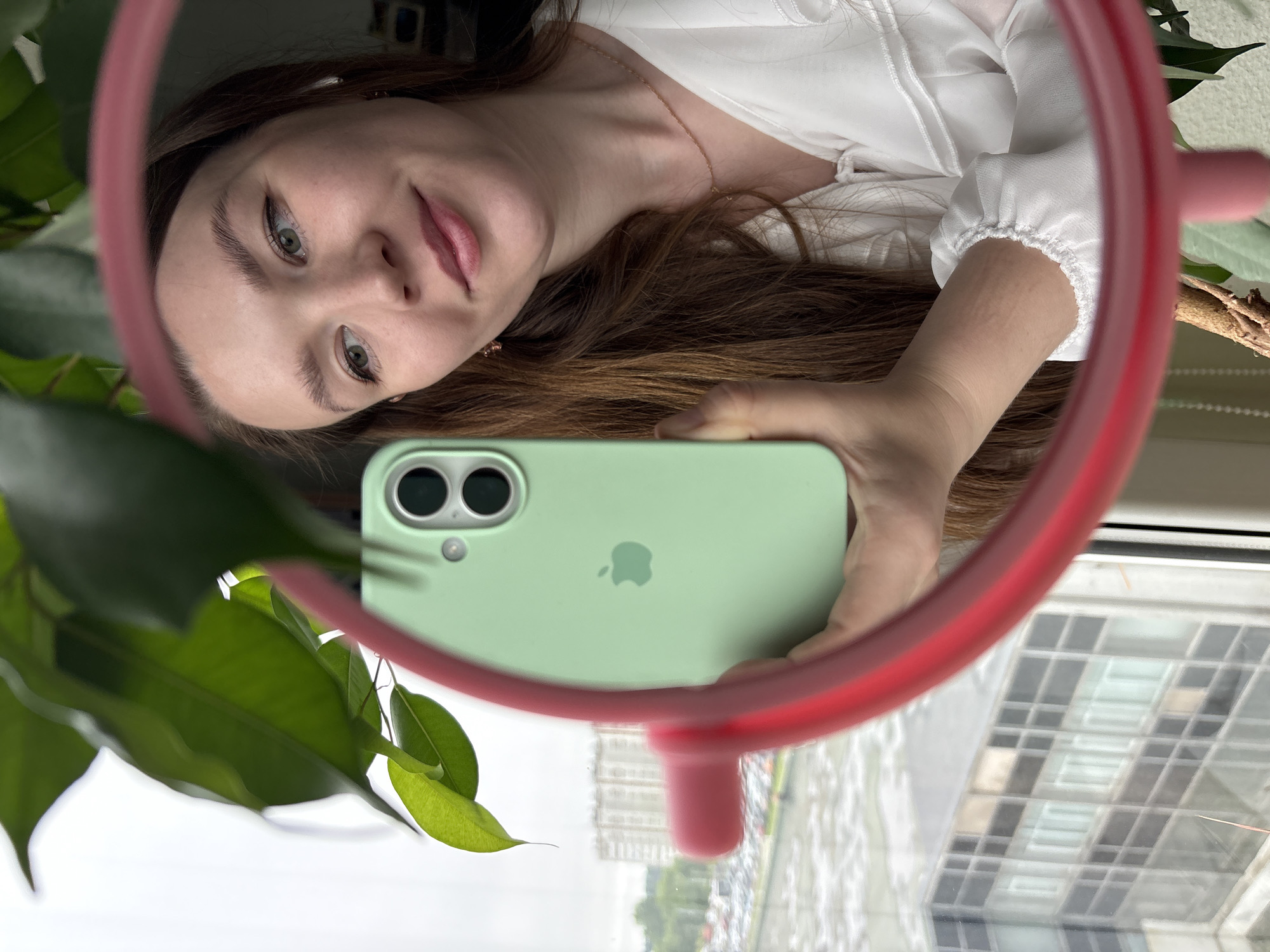 The image is a mirror selfie featuring a young woman reflected in a round mirror with a thick pink or red frame. The woman, seen from the chest up and appearing upside down due to the reflection, has long brown hair, fair skin, and light-colored eyes. She wears a white, possibly ruffled, blouse and a delicate gold chain necklace, and sports a faint, pleasant smile.
She holds a light green smartphone, confirmed as an iPhone by its visible Apple logo and dual camera system, pointing it towards the mirror to capture the shot. Her hand and arm, adorned with the gathered elastic cuff of her white sleeve, are also visible.
The scene is indoors, likely near a window, with a vibrant green-leafed plant partially obscuring the left side of the mirror and the outside view. The background, reflected in the lower portion of the mirror and glimpsed through the window, reveals an urban landscape under a bright, possibly overcast, daytime sky. Tall buildings with distinctive window patterns and a street or parking area with vehicles are visible, suggesting a city environment. The setting is located in Izhevsk, Russia.