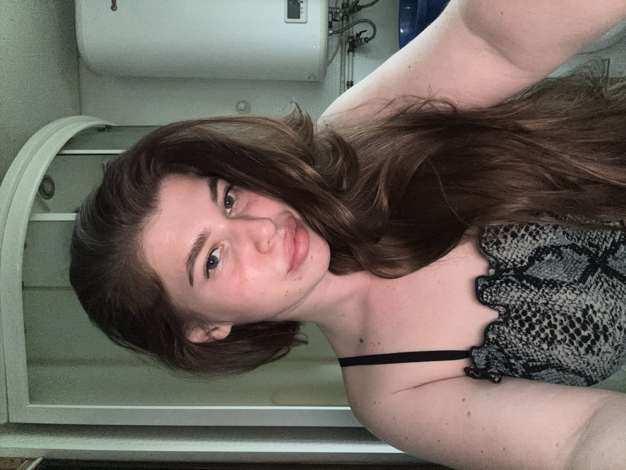 A young woman is facing the camera with a slightly tilted head. She has long, brown hair and light skin with some visible freckles. She is wearing a snakeskin-patterned top with a black strap. The background appears to be a bathroom, with a white water heater, pipes, and a shower door visible. The location is not evident from the image, so identifying the city of Мещёры, Russia is not possible.
