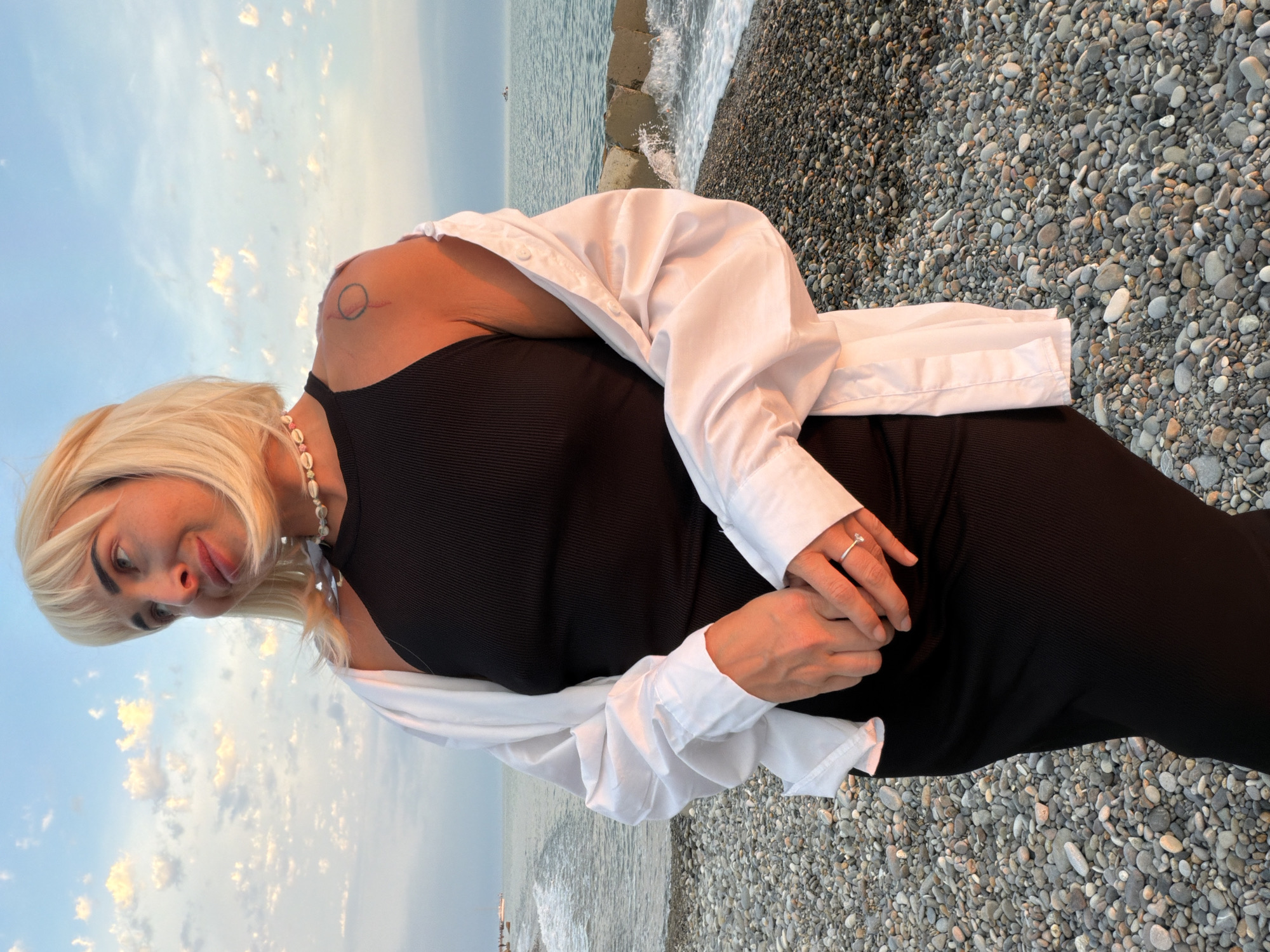 In Sochi, Russia, a woman with blonde bobbed hair and bangs stands on a pebble beach at dusk. She is wearing a black, form-fitting ribbed dress, a white unbuttoned shirt draped over her shoulders, and a shell necklace. Her hands are clasped in front of her, and she wears a ring on her left hand. A tattoo is visible on her right shoulder. The sea with gentle waves is behind her, with a breakwater in the distance. The sky is a blend of blue and scattered clouds, illuminated by the setting sun, casting a warm orange glow on the scene.