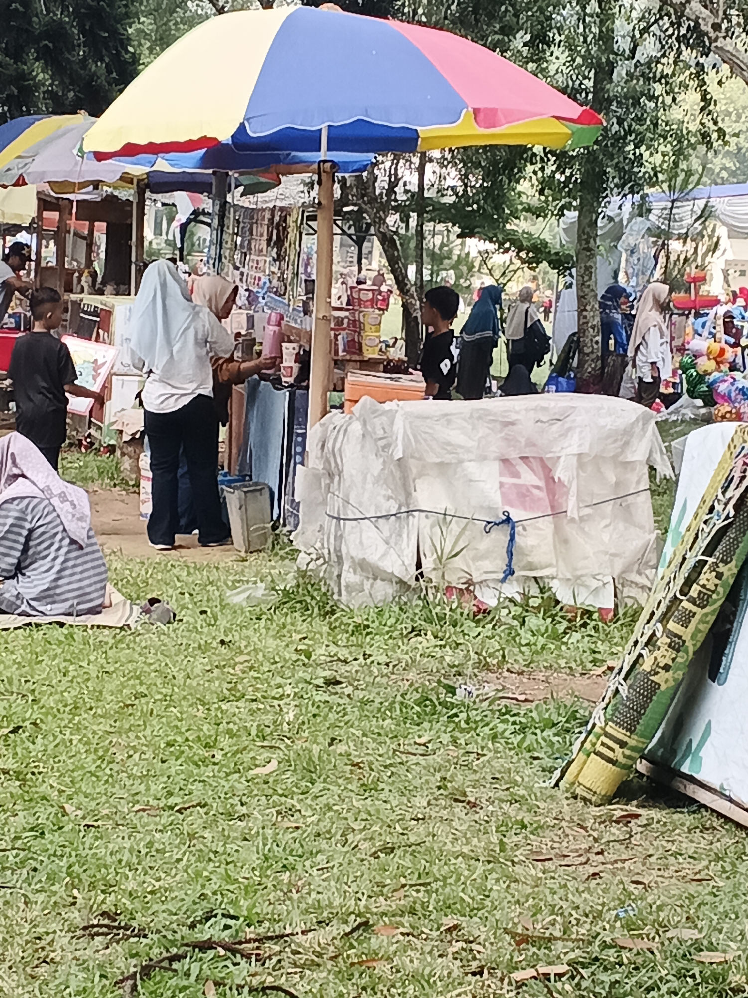 The image depicts an outdoor market scene in Perbawati, Indonesia, likely set up on a grassy area with trees in the background. A large, colorful umbrella, divided into segments of yellow, blue, red, and green, is a prominent feature, providing shade over a stall. Beneath the umbrella, several individuals are visible, engaged in various activities. Two young people, one in a black t-shirt and the other in a white hijab, stand at a stall, possibly interacting with a vendor. Another person in a black hijab is also present near the stall. In the foreground, to the left, a person in a striped grey and white hijab is seated on the grass. To the right, a white tarp-covered structure is visible, with a blue rope tied around it. Several items are displayed at the market stalls, including what appear to be books or packaged goods. The ground is covered with green grass, interspersed with fallen leaves and twigs. A portion of a patterned textile, possibly a mat or rug, leans against a light-colored structure on the far right. The overall atmosphere suggests a casual, bustling market environment.
