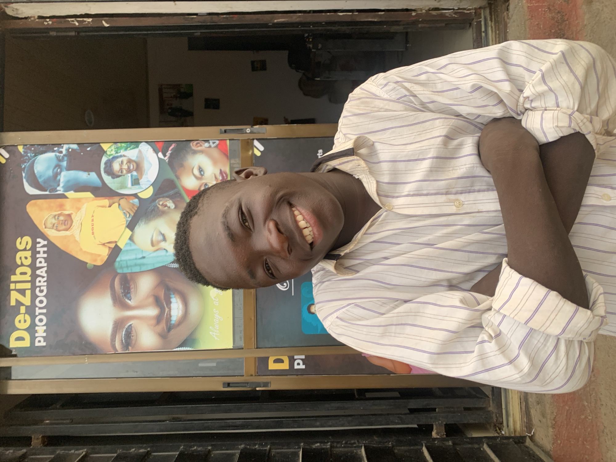 A young Black man is posing in front of a photography studio named "De-Zibas Photography". He is smiling with his arms crossed. He is wearing a white long-sleeved shirt with light purple stripes. The photography studio's advertisement displays various photographs of people. The location is the city of Jos, Nigeria.