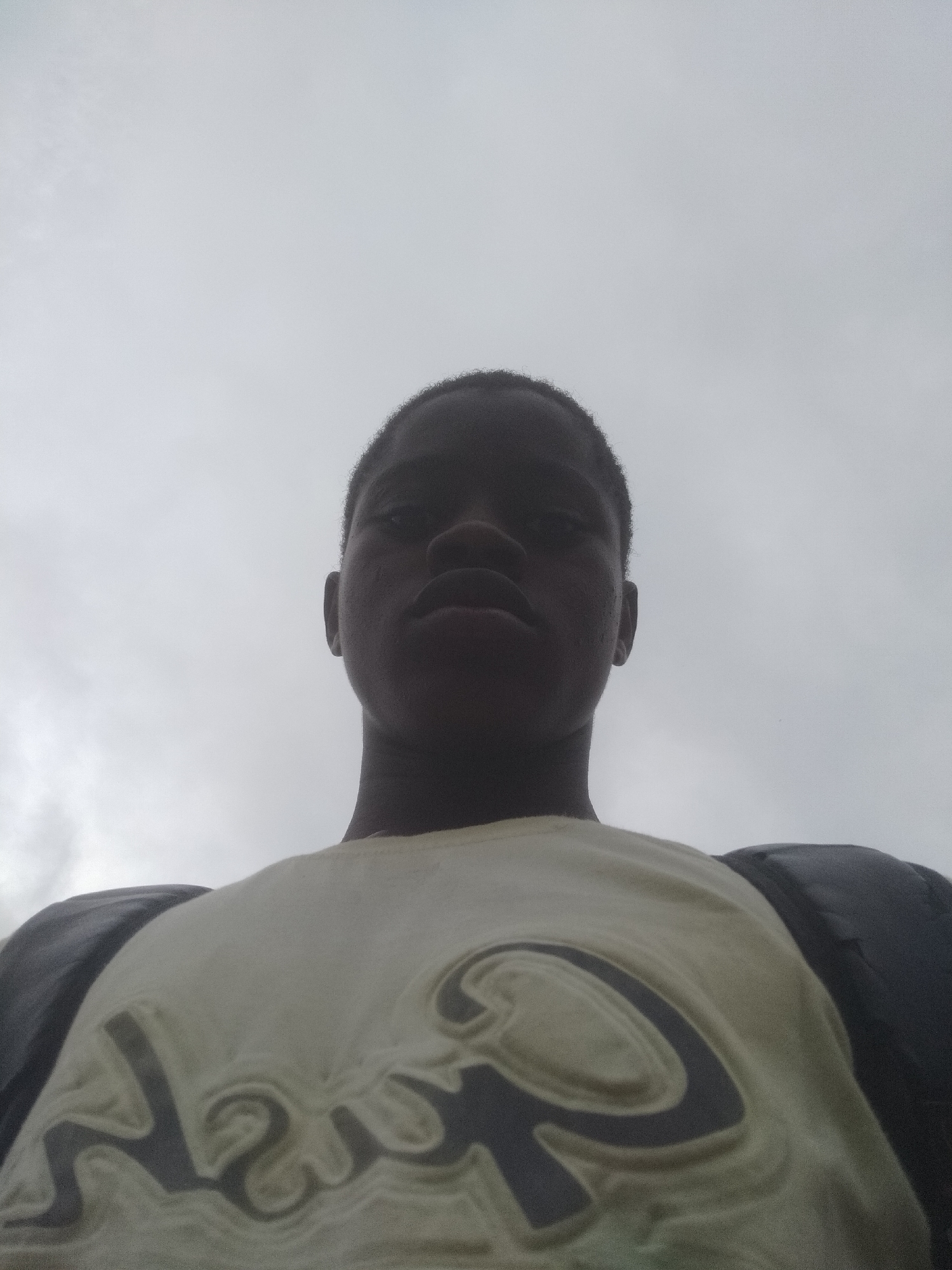 A low-angle headshot shows a young, dark-skinned male. He is wearing a light-colored t-shirt with raised, indistinct dark lettering. The background is a cloudy sky. The city is unknown.