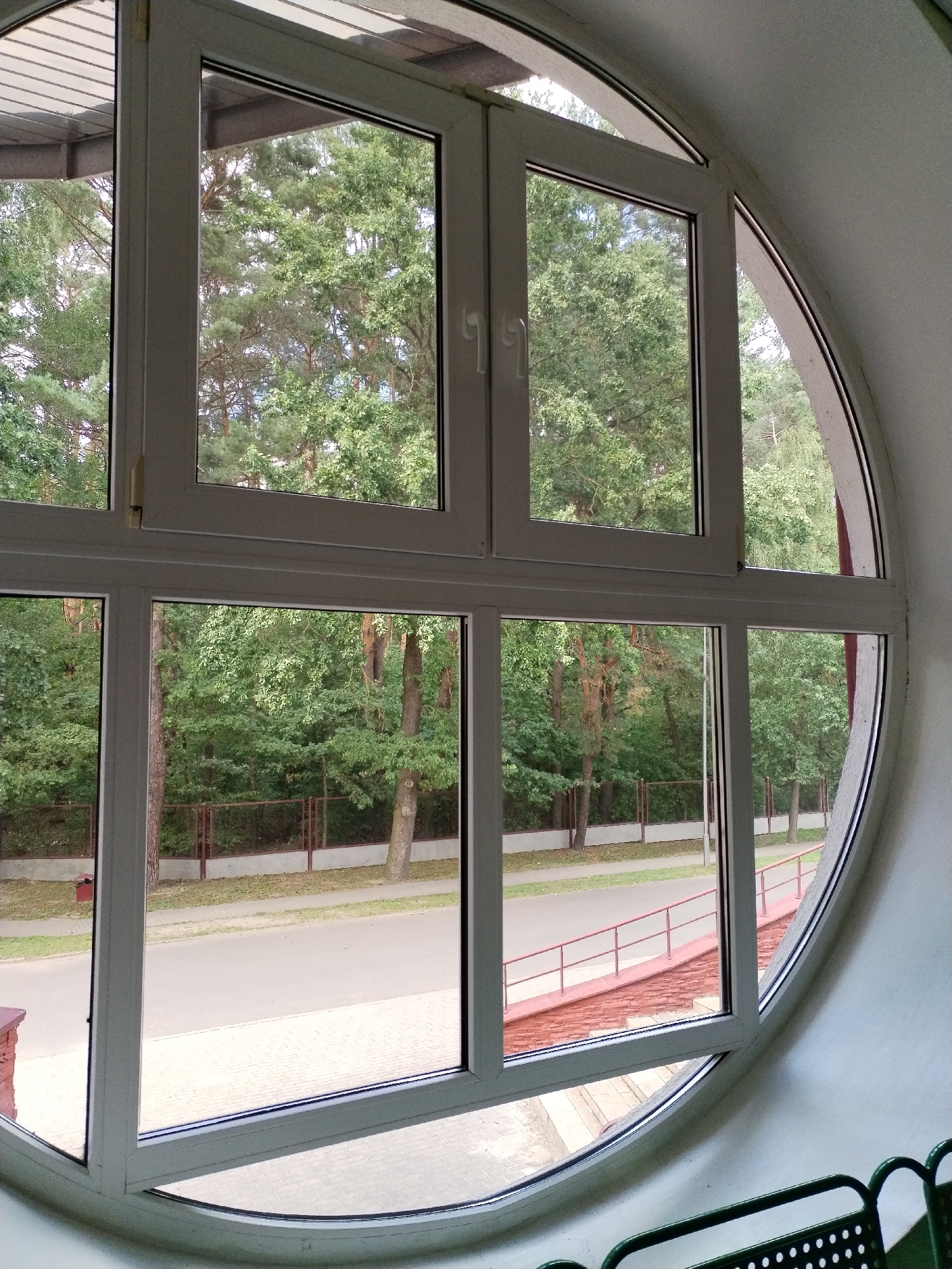The image shows a large, circular window with a white frame, divided into several panes. Through the window, a view of lush green trees and a paved road is visible. The trees appear to be a mix of deciduous and coniferous species, and they form a dense canopy. A fence runs along the edge of the road, and beyond that, more trees can be seen. The road itself is asphalt, and there are some bushes and a railing visible on the side. The overall impression is that of a natural, somewhat rural setting, possibly a park or a wooded area. The window is looking out from an interior space, as suggested by the white interior wall and the edge of a green metal chair visible in the foreground. There are no people visible in the image. The specific location is Хідрынскі сельскі Савет, Belarus, which is a rural administrative division within Belarus, known for its natural landscapes and agricultural activities.