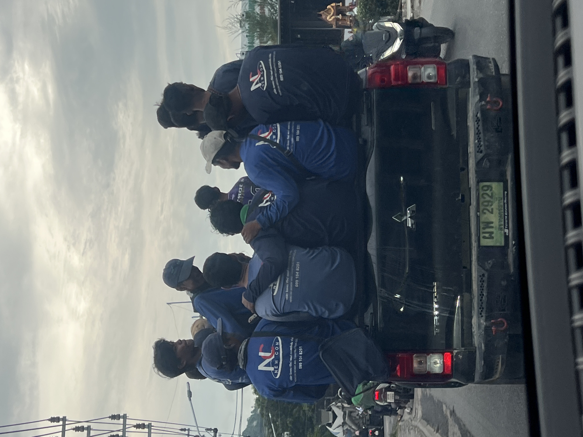 The image shows a group of men, likely workers, being transported in the open bed of a black Mitsubishi pickup truck on what appears to be a road. The sky is overcast, indicating daytime. The men are dressed in matching long-sleeved blue or navy shirts, many wearing caps or hats, and are crowded together, standing and possibly sitting.
Notable details provide context about the scene and location. The shirts feature a prominent logo that reads "NEW CON," with smaller text identifying it as "DESIGN AND CONSTRUCTION CO.,LTD." and phone numbers such as "099 104 8251" and "099 104 8259," suggesting they are employees of a construction or design company. The pickup truck's license plate displays Thai script "ผพ 2929," indicating the location is Thailand. A "TOYOTA GAZOO Racing" sticker is also visible on the tailgate, along with the "Mitsubishi" brand name.
In the background, utility poles with numerous wires are visible on the left, and other vehicles, including motorcycles, are present on the road. Distant foliage and structures, including what appears to be a golden spire (possibly part of a temple), contribute to the semi-urban setting. The activity depicted is a routine commute or transportation of workers.