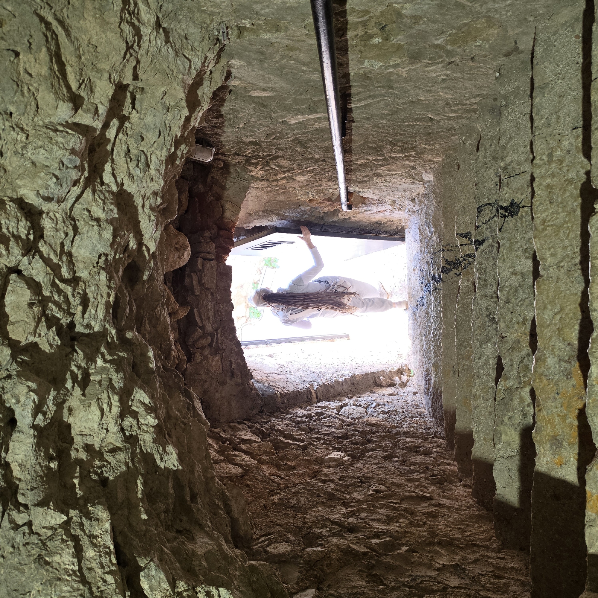 The image shows a long, narrow, man-made stone tunnel or cave. The walls are rough, irregular, and light brown, with some darker areas due to shadow. The floor of the tunnel is also made of stone and appears uneven. At the far end of the tunnel is a bright, open space that appears to be outdoors, as some green vegetation is visible. In this open space, a person wearing light-colored clothing is visible, with their face obscured by the brightness. They appear to be reaching up with one hand. A metal bar is fixed across the top of the tunnel near the opening, suggesting a possible support structure or installation. The location is in the city of Unknown, Unknown.