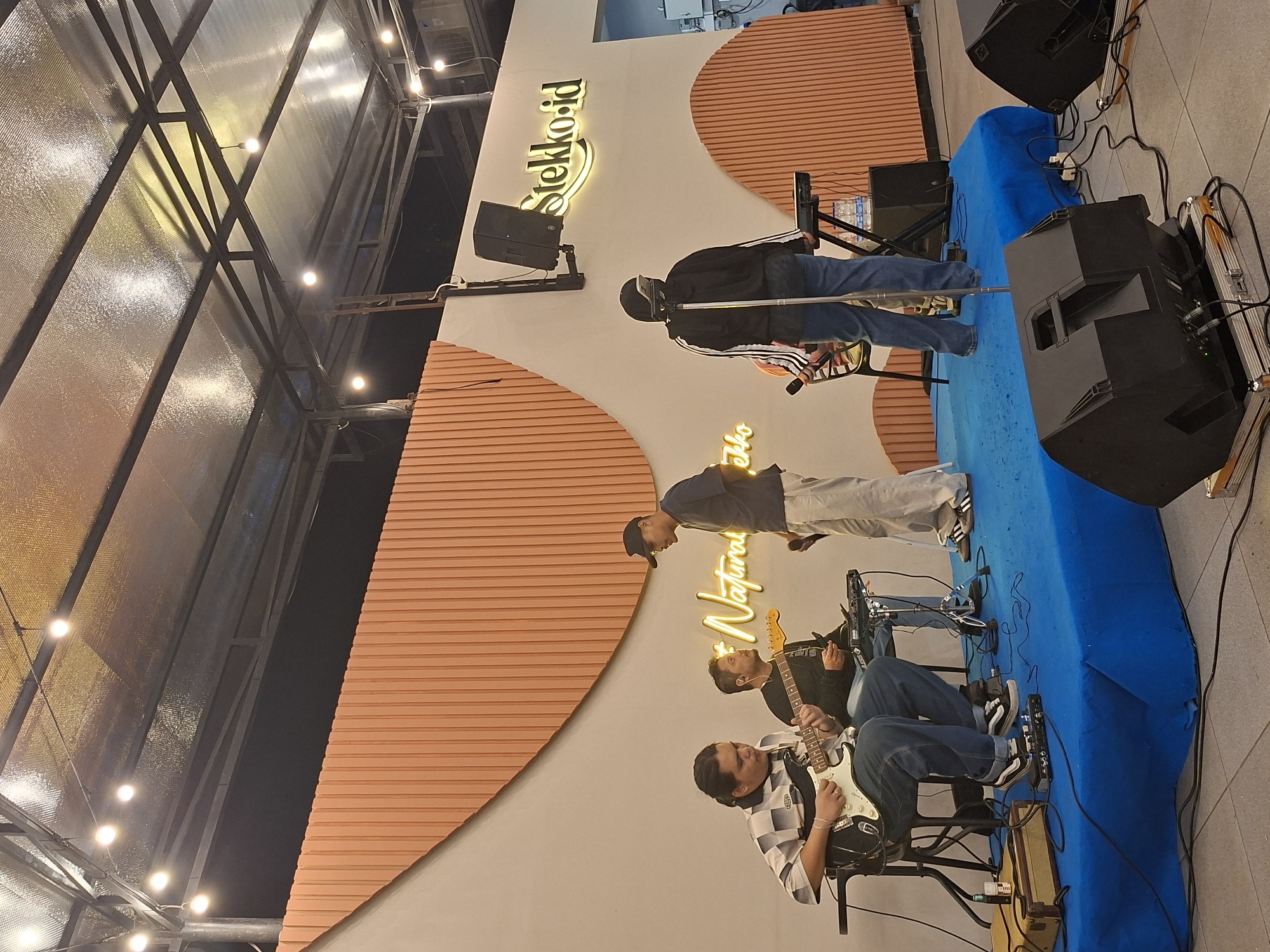 The image shows a band performing on a stage in Lhokseumawe, Indonesia. The stage is covered in a blue cloth. Two guitarists are seated, and a vocalist is standing behind them. A second person with a microphone stand is standing off to the side, and another individual is standing behind the vocalist. Two large black speakers flank the stage. Behind the band is a wall decorated with horizontal orange stripes arranged in a semi-circular pattern. The word "Stekko.id" is displayed on the wall in illuminated letters. Above the scene is a metal framework and lighting fixtures, suggesting an open-air or partially covered venue.