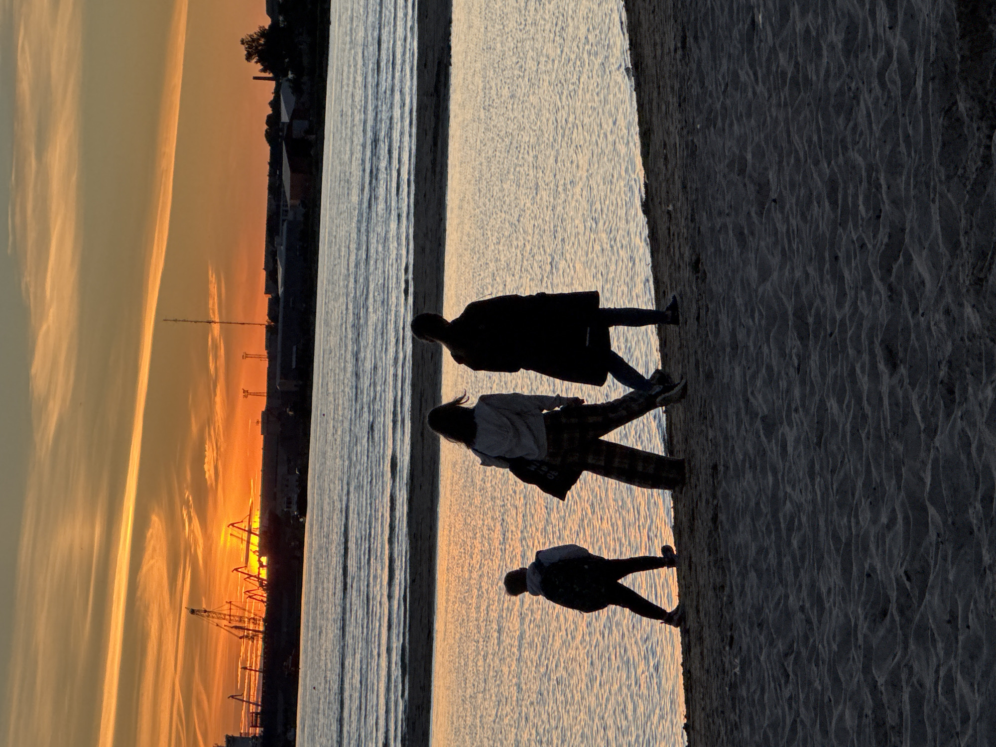 The image depicts a sunset over the Baltic Sea at the Port of Tallinn, Estonia. In the foreground, three figures, silhouetted against the water and sky, walk along a sandy beach. The two adult figures are walking side-by-side, with the figure on the right wearing a dark coat and the figure on the left wearing a light-colored hooded sweatshirt and checkered pants. A child, carrying a backpack, walks slightly ahead of them. The water is calm with gentle ripples reflecting the warm hues of the sunset. In the background, the port is visible with several cranes and industrial buildings silhouetted against the vibrant orange and yellow sky. The sun is low on the horizon, casting a warm glow over the scene.