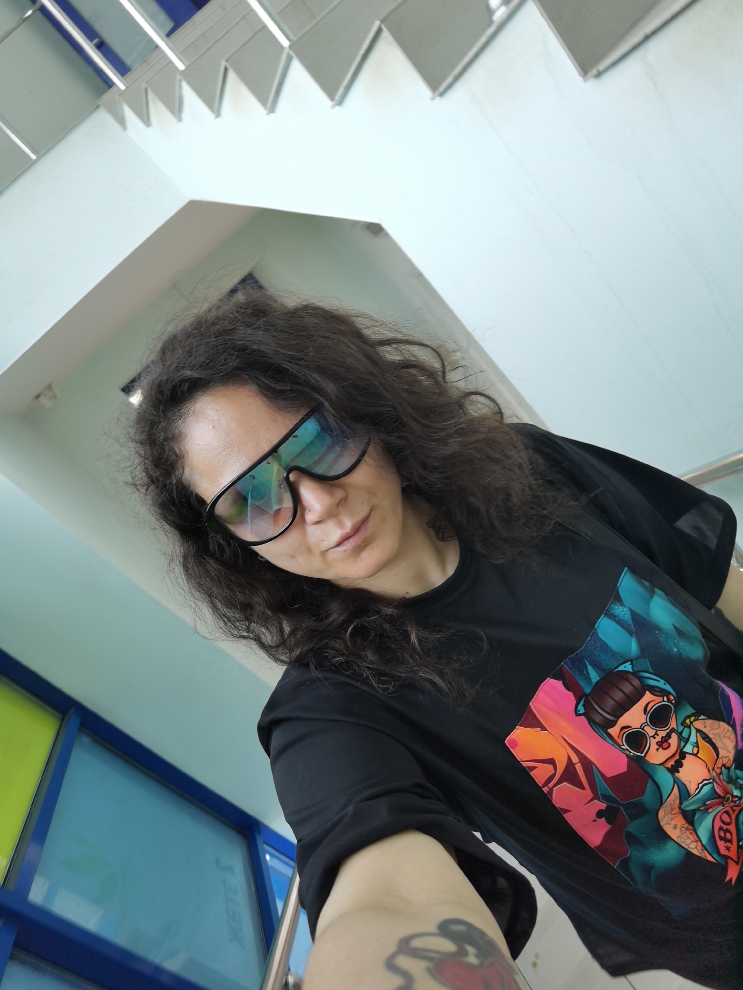 A medium shot photo shows a woman with dark curly hair and large, reflective sunglasses. She is wearing a black t-shirt with a colorful graphic print of a pin-up style character. The woman is looking down and to the left, and her right arm is extended towards the camera, showing a tattoo on her forearm. The background consists of a light blue wall and a ceiling with angular, white elements. There are also some blue and green framed windows visible in the lower left corner. The scene appears to be indoors, possibly in a public space like a waiting area or transit hub. The specific location in Chaikovsky, Russia, cannot be determined from the visual information provided in the image.