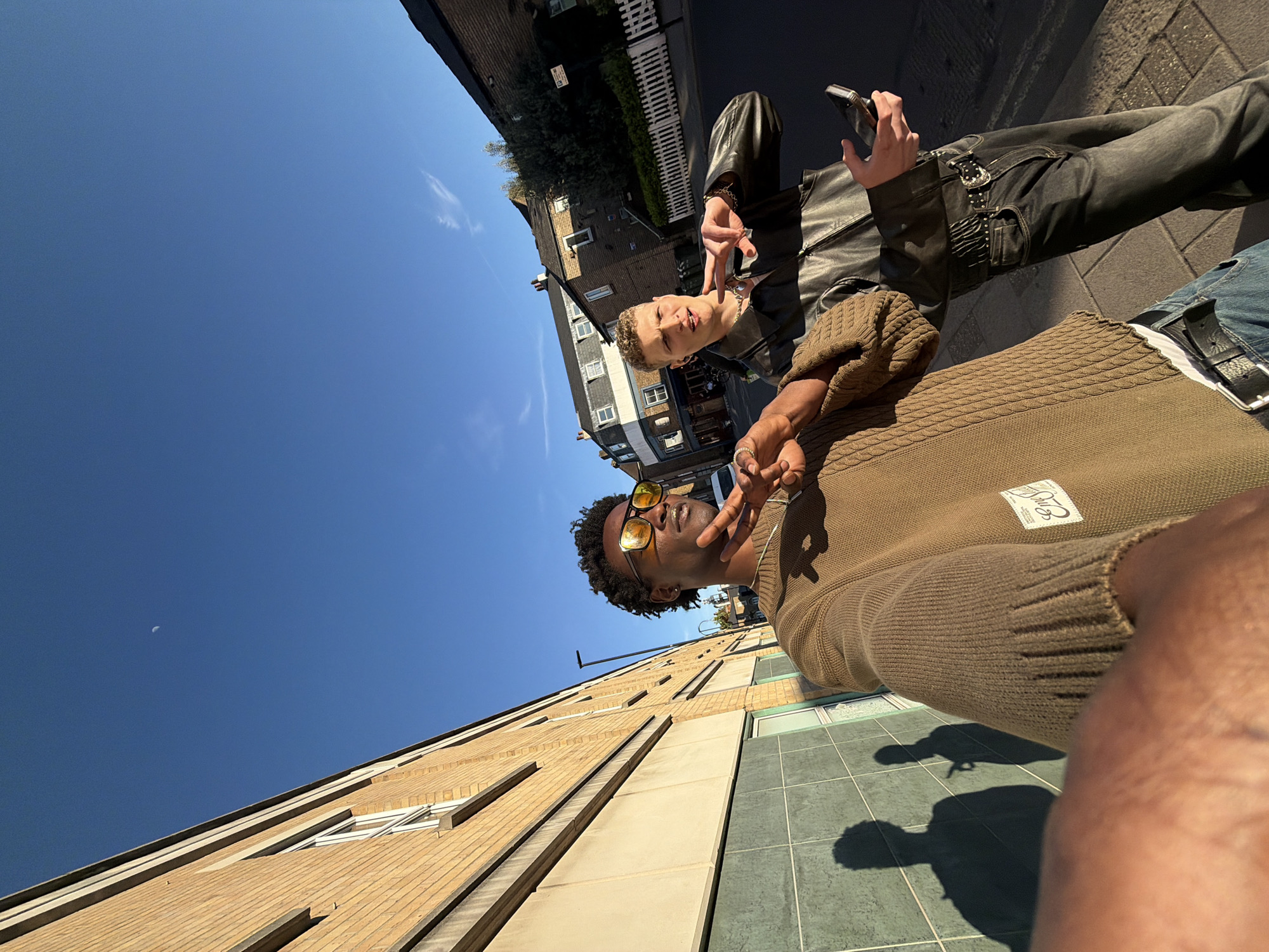 This dynamic image, taken at a low angle and rotated clockwise, captures two young men posing on a sunny street, likely in London, United Kingdom, given the architectural style. The perspective makes the ground appear as a side wall and the sky as the top-right quadrant.
In the foreground, a man with dark curly hair and dark sunglasses with yellow lenses wears a brown cable-knit sweater. He flashes a peace sign with his right hand. On his sweater, a rectangular patch with visible text reads "St. Gregory & Co." and "Est. 1956" or similar. Parts of his arm are visible in the bottom right, suggesting he might be holding the camera for a selfie.
Standing slightly behind and to his left is a man with short, light-colored curly hair. He is dressed in a black leather jacket, dark jeans, and an ornate silver belt buckle. He also makes a peace sign with his left hand while holding a smartphone in his right. He has a casual, slightly smiling expression.
The background reveals typical urban architecture: brick buildings with multiple windows, some featuring white fences, and trees/bushes. The clear blue sky dominates much of the upper frame, showcasing a distinct crescent moon and a few wispy clouds. Bright sunlight casts long, clear shadows of the two individuals onto the tiled pavement below, indicating a bright daytime setting, possibly morning or late afternoon. The overall mood is casual and friendly, capturing a moment between friends on a clear day.