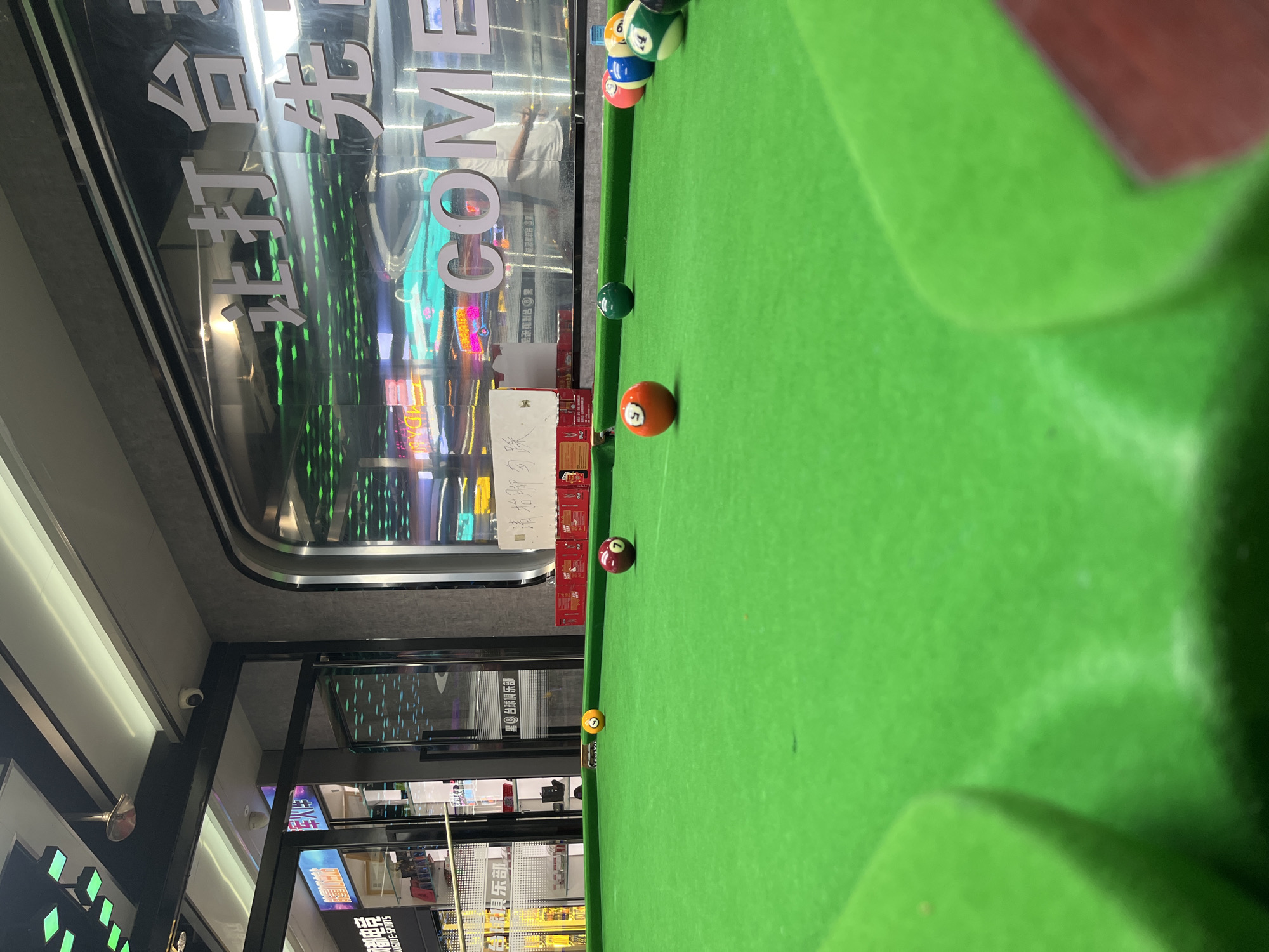 This is a tilted, medium-shot photograph taken at a billiards hall in Daxing District, China. The green felt of a pool table dominates the foreground and right side of the image. Several billiard balls are scattered across the table. One ball, colored red with a white number '5' on it, is closest to the viewer. Further up the table, a solid green ball is visible, followed by a cluster of balls, including a red '7' and a yellow '13'.
In the background, through a large glass window, a store interior is visible, illuminated by neon lights. Some of the text on the window is legible due to the reflection: "COME" and Chinese characters that appear to say "台球俱乐部" (Billiards Club). The overall lighting suggests it is nighttime or indoors with artificial lighting.
The scene appears to be set up for a game of pool. There are no people clearly visible actively playing, but a person in a white shirt can be seen in the reflection within the glass window. The atmosphere seems casual and recreational. The floor in the background is tiled, and shelving units are visible, possibly stocked with merchandise. A security camera is mounted on the ceiling in the upper left corner of the image.