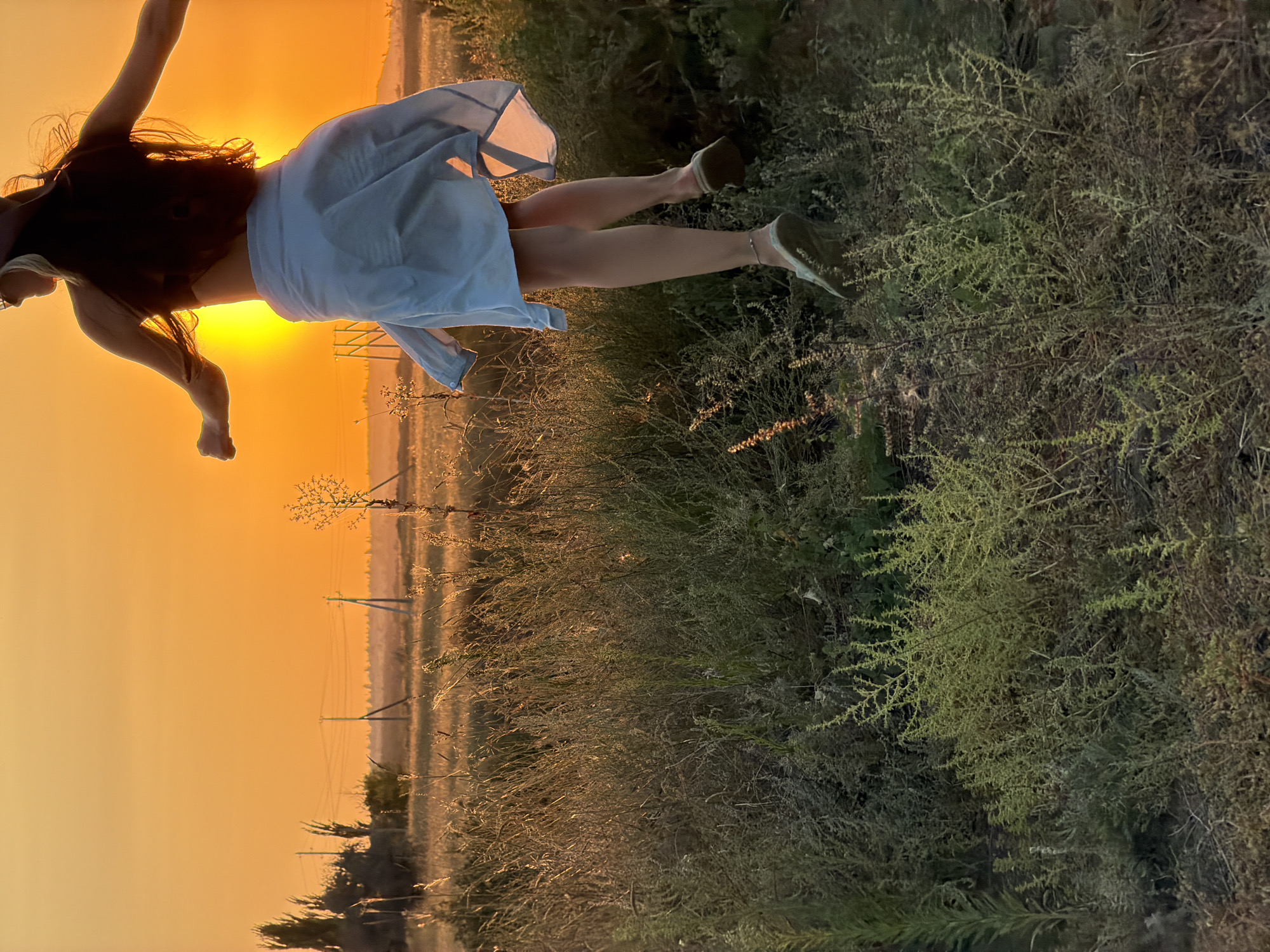The image depicts a woman in mid-air, jumping in a field during sunset. She is wearing a light-colored skirt and a dark top, with her back to the camera. Her arms are outstretched, and her hair is flowing. The sun is setting behind her, casting a warm orange glow over the scene. The background shows a field of dry grass and some trees. Power lines and poles are visible in the distance, suggesting a rural or semi-rural setting. The lower portion of the image is dominated by tall, dry grass and other vegetation. The overall impression is one of freedom and joy, captured in a beautiful natural light.