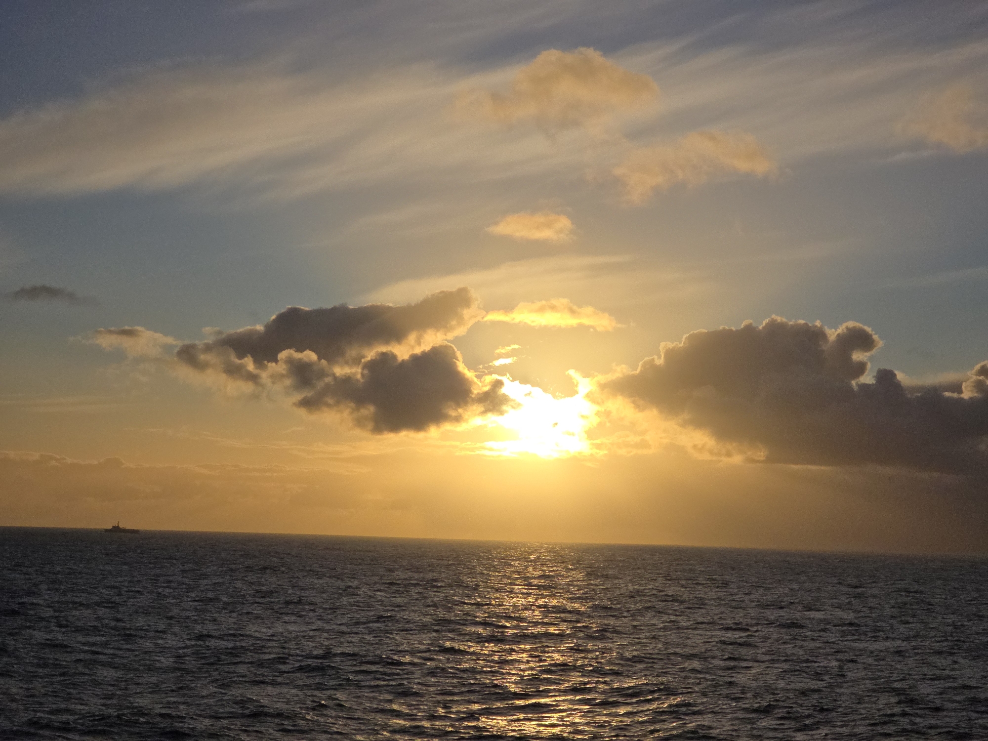 The image depicts a sunset over the ocean. The sky is a mix of blue, gray, and yellow hues, with clouds obscuring the sun. The sun's rays create a bright glare where it peeks through the clouds, casting a golden glow on the water. The water appears dark with lighter reflections indicating waves. A small silhouette of a ship is visible on the horizon. The location is over the ocean in the city of Unknown, Unknown.
