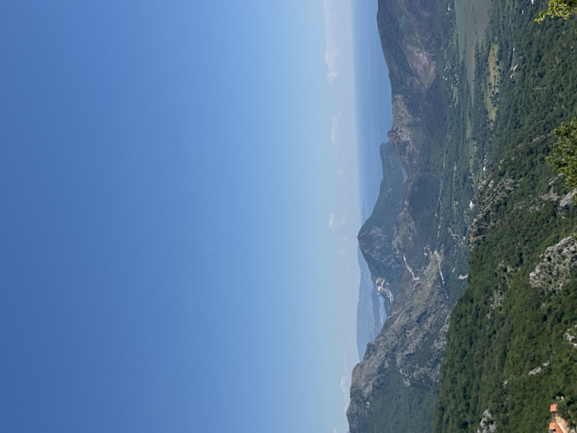 A landscape photograph, taken from an elevated perspective, features a mountainous terrain under a clear blue sky. The mountains, primarily covered in dense green vegetation, descend into a valley where small villages or settlements, possibly including Novoselje, Montenegro, are interspersed with patches of greenery. The peaks are rugged, showcasing rocky textures and steep inclines. A body of water, possibly the sea or a large lake, is faintly visible in the background, partially obscured by the atmosphere or a slight haze. The sky dominates the left side of the frame, its color a gradient of azure blue.
