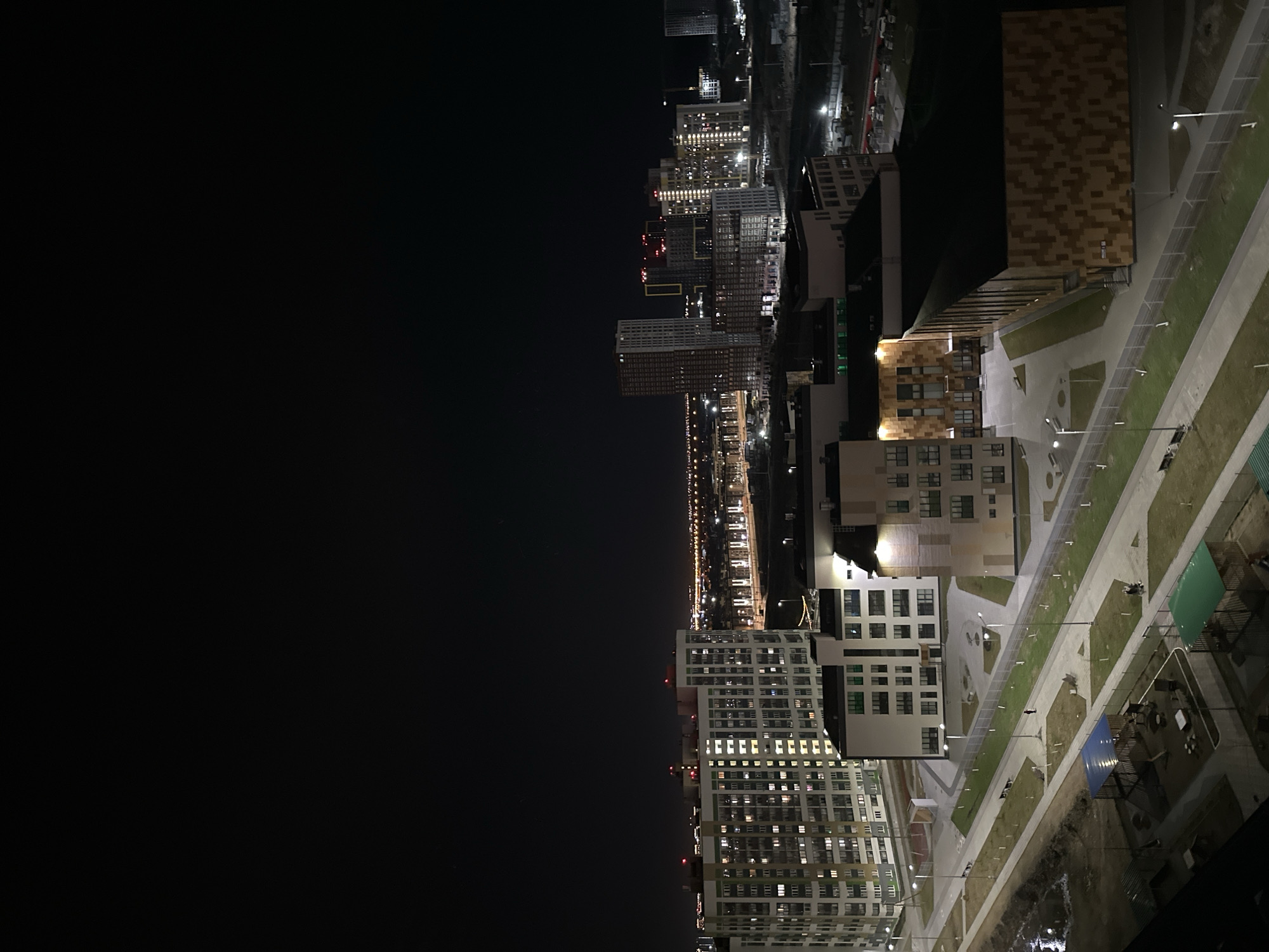 A night aerial shot of a residential area in Yekaterinburg, Russia, shows a mix of high-rise apartment buildings and lower-rise brick structures along a wide boulevard lit by streetlights. The buildings' illuminated windows reveal a vibrant urban living environment. The left portion of the image is dominated by a dark sky. The scene includes green spaces, sidewalks, and modern architectural designs.